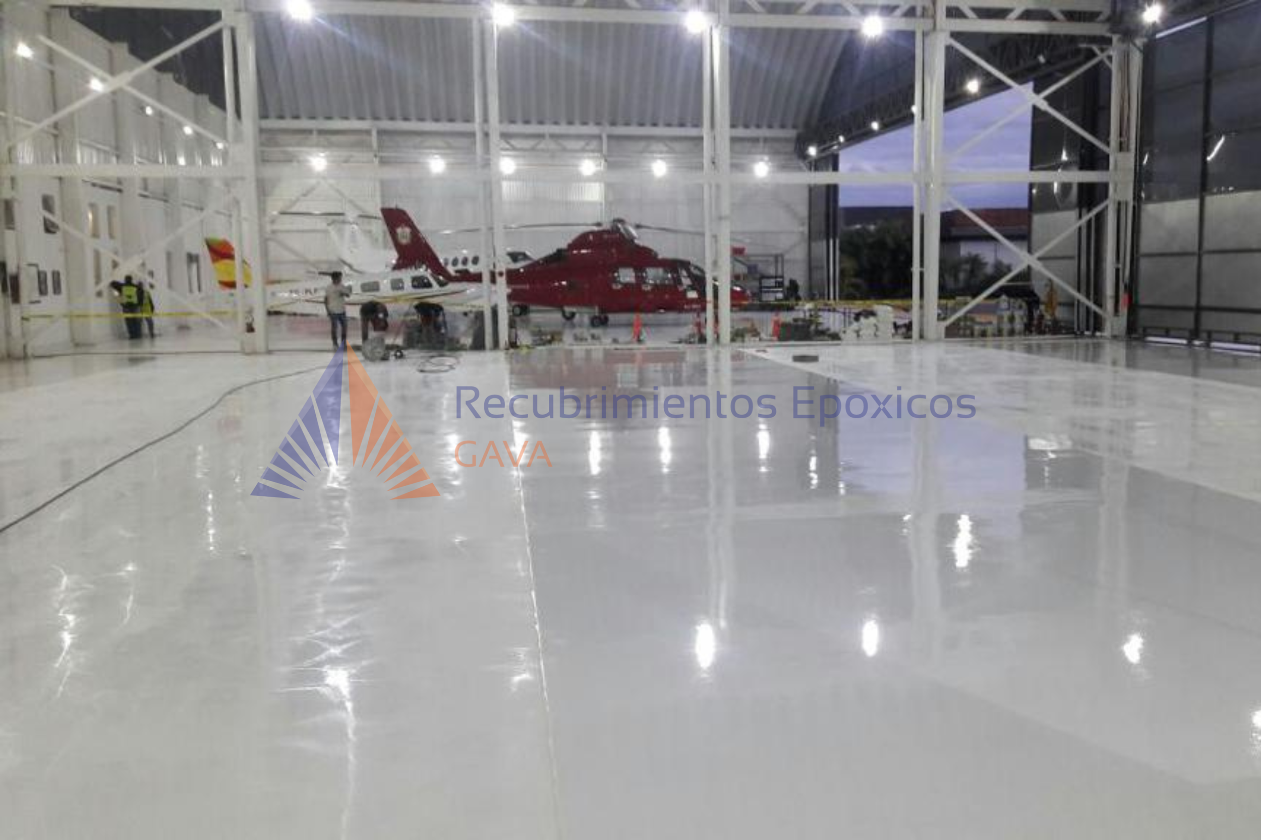 Interior de un hangar con aviones de diferentes modelos, uno de ellos de color rojo, y personal técnico trabajando. Hay marcas de recubrimiento epóxico en el suelo y la estructura del hangar está pintada de blanco.