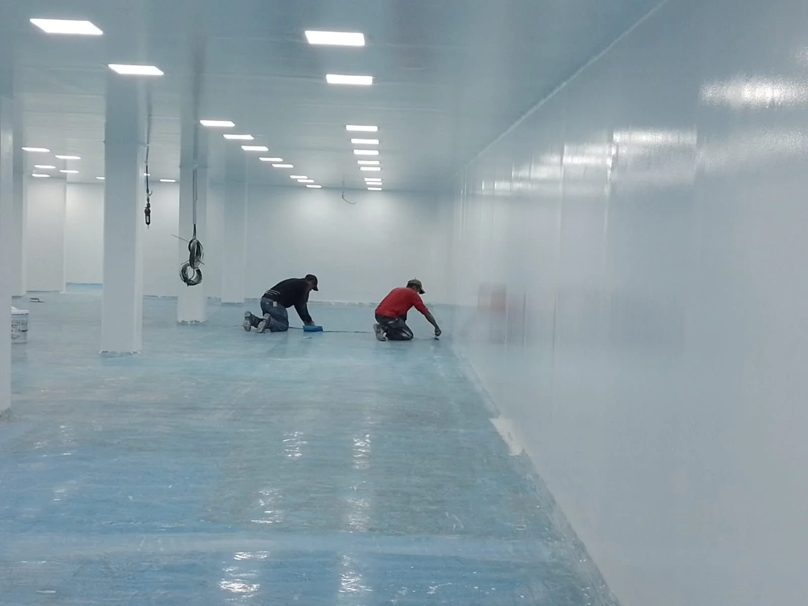 Dos trabajadores aplicando pintura en el suelo de una gran instalación con paredes y techo blancos, iluminación LED en el techo.