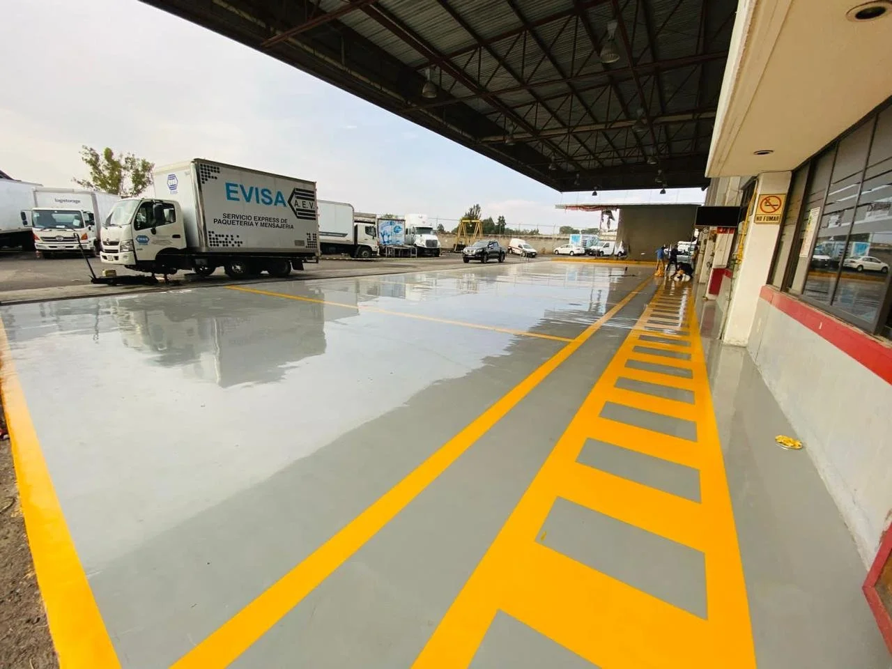 Zona de estacionamiento en un área industrial con suelo recién pintado en gris y líneas de demarcación en amarillo. Hay varias camionetas y vehículos en el fondo, y un techo que cubre parcialmente la estructura. A la derecha, ventanas y un cartel de 'No Fumar'.