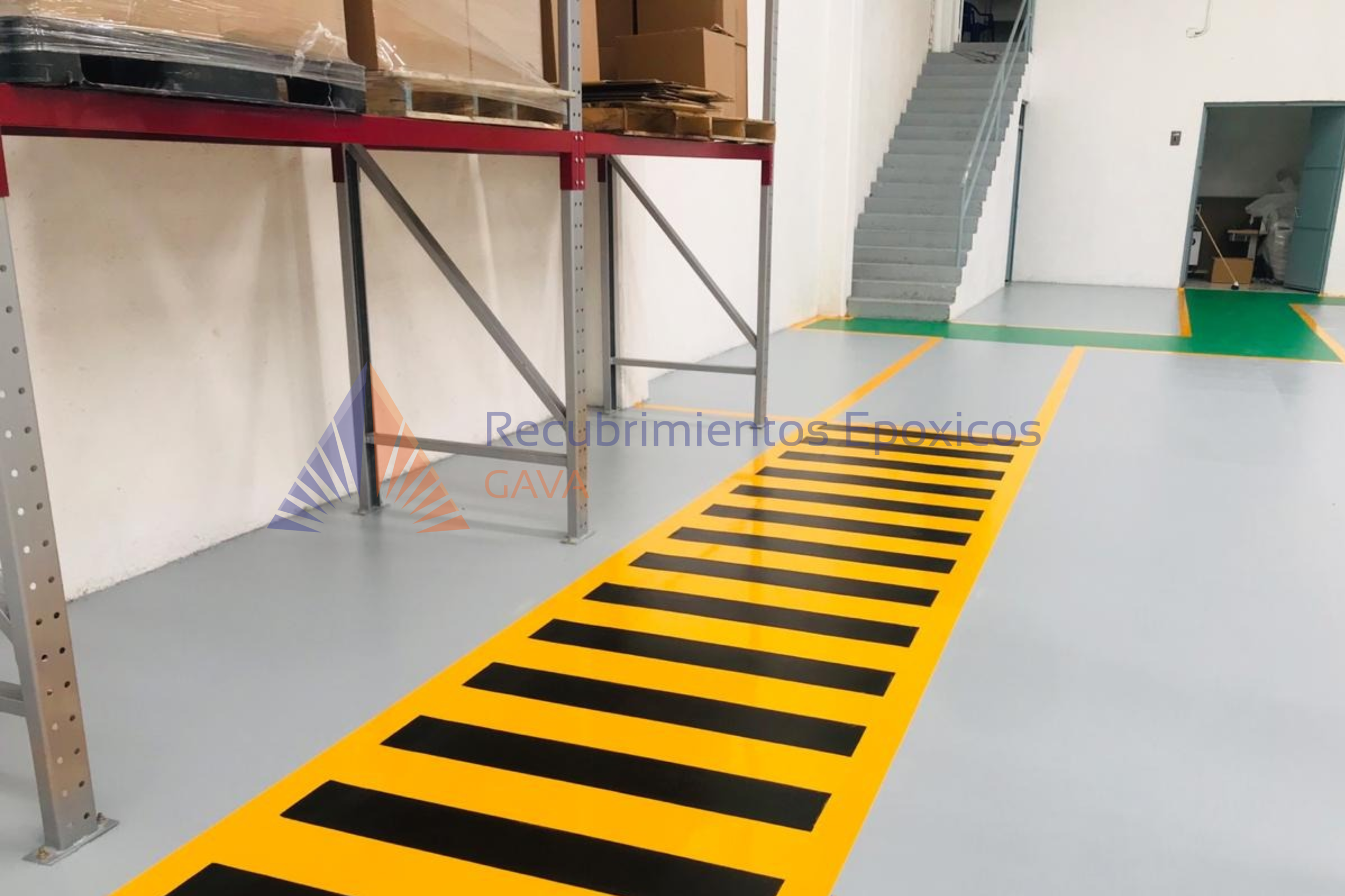 Piso de color gris con líneas amarillas y negras en el suelo, estructura metálica en el lado izquierdo, escaleras de concreto y pasamanos en el fondo, un espacio interior de almacén o fábrica con paredes blancas y zona de entrada o salida en la esqui