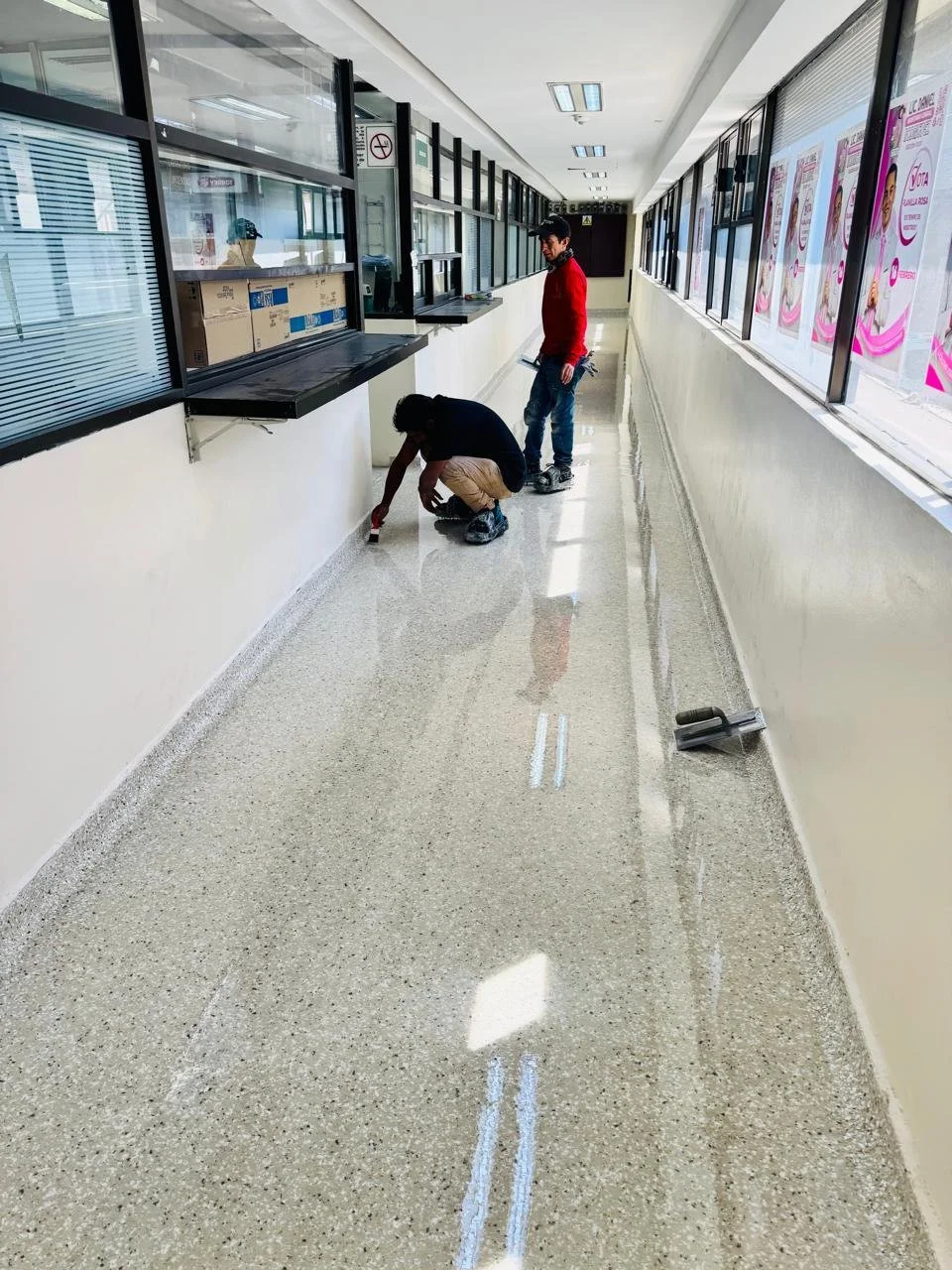 Dos personas trabajando en la reparación del suelo en un pasillo de un edificio, uno está pintando o limpiando, y el otro observa, con ventanas y publicidad en el fondo.