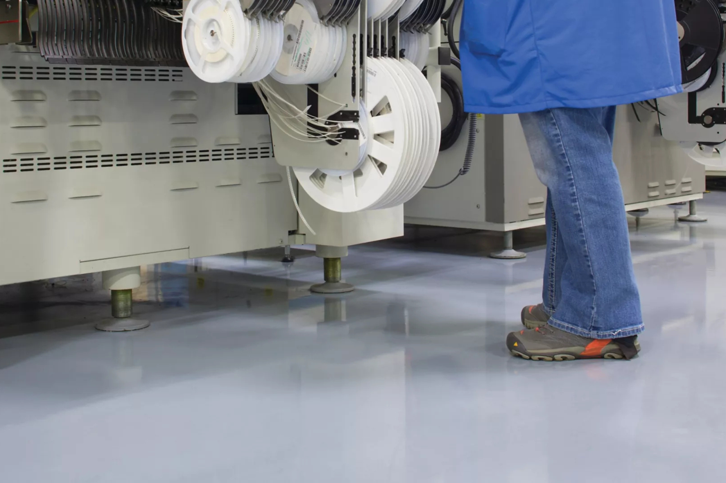 Persona de pie usando ropa azul, jeans y zapatos deportivos, trabajando con bobinas de cable blanco en un entorno industrial o de fabricación.