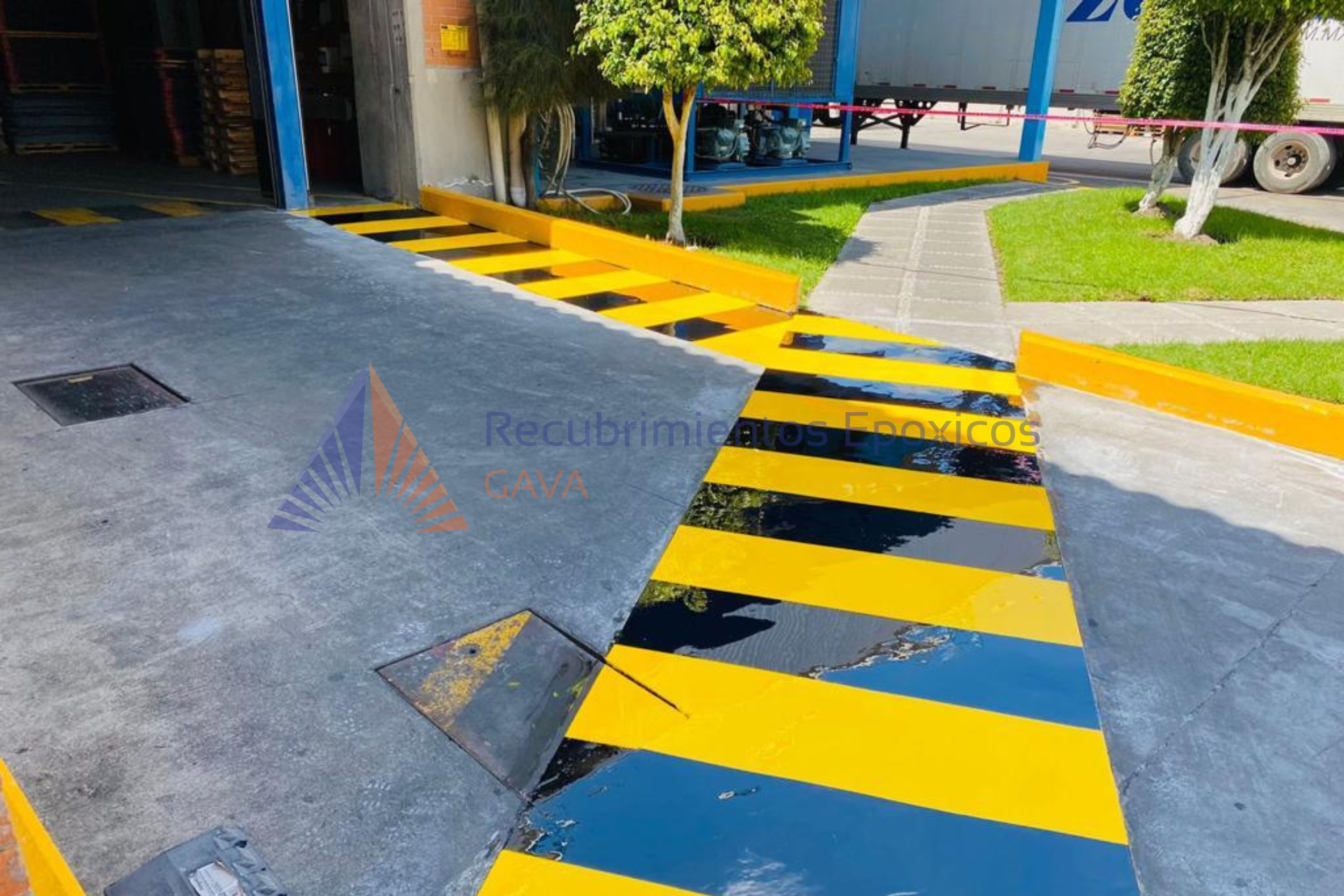 Reborde de seguridad amarillo y negro en entrada de almacén con pasillo de cemento y césped con árboles y camino de loseta.