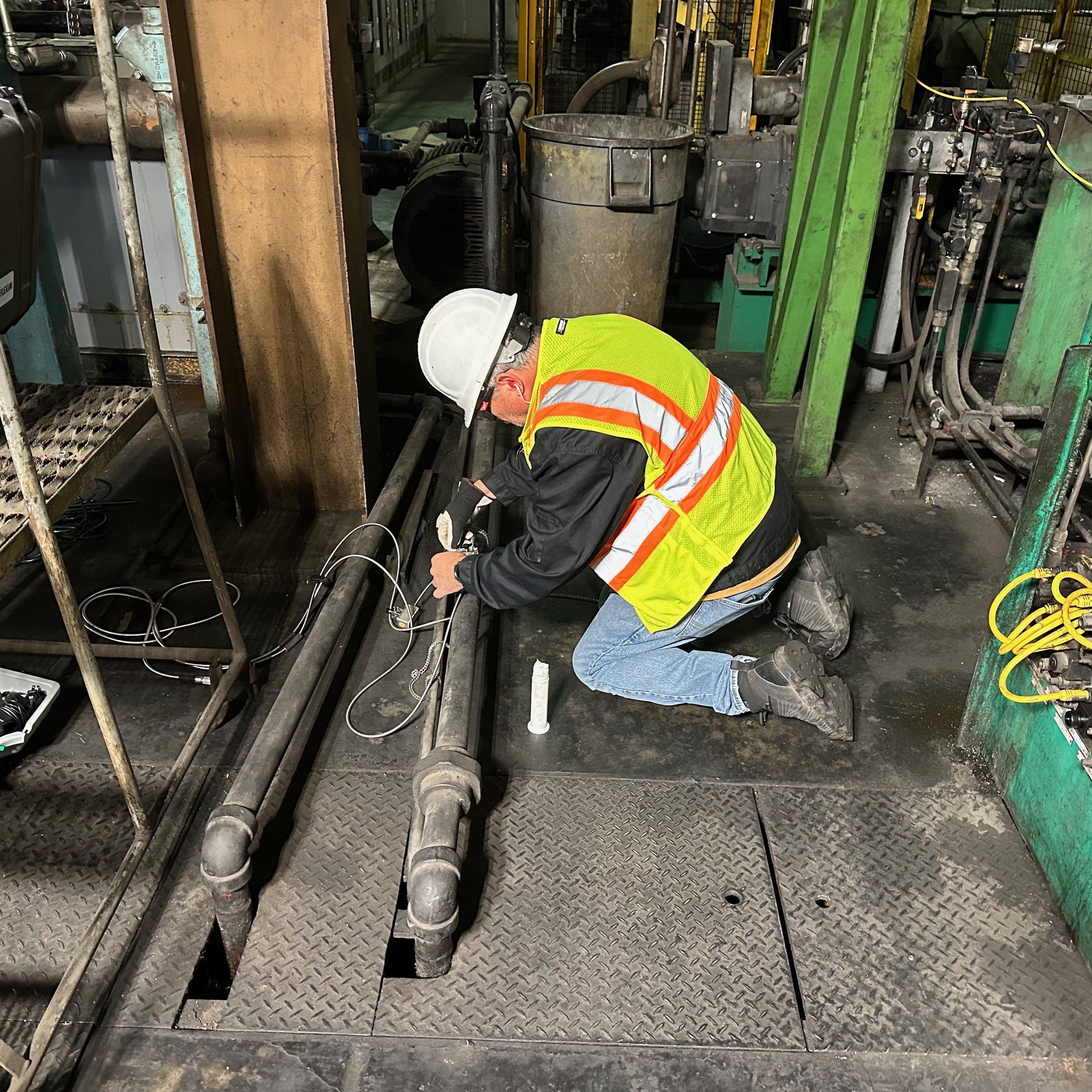 A worker in a white helmet, yellow safety vest, and black jacket kneeling on a metal floor, working on large pipes in an industrial setting.