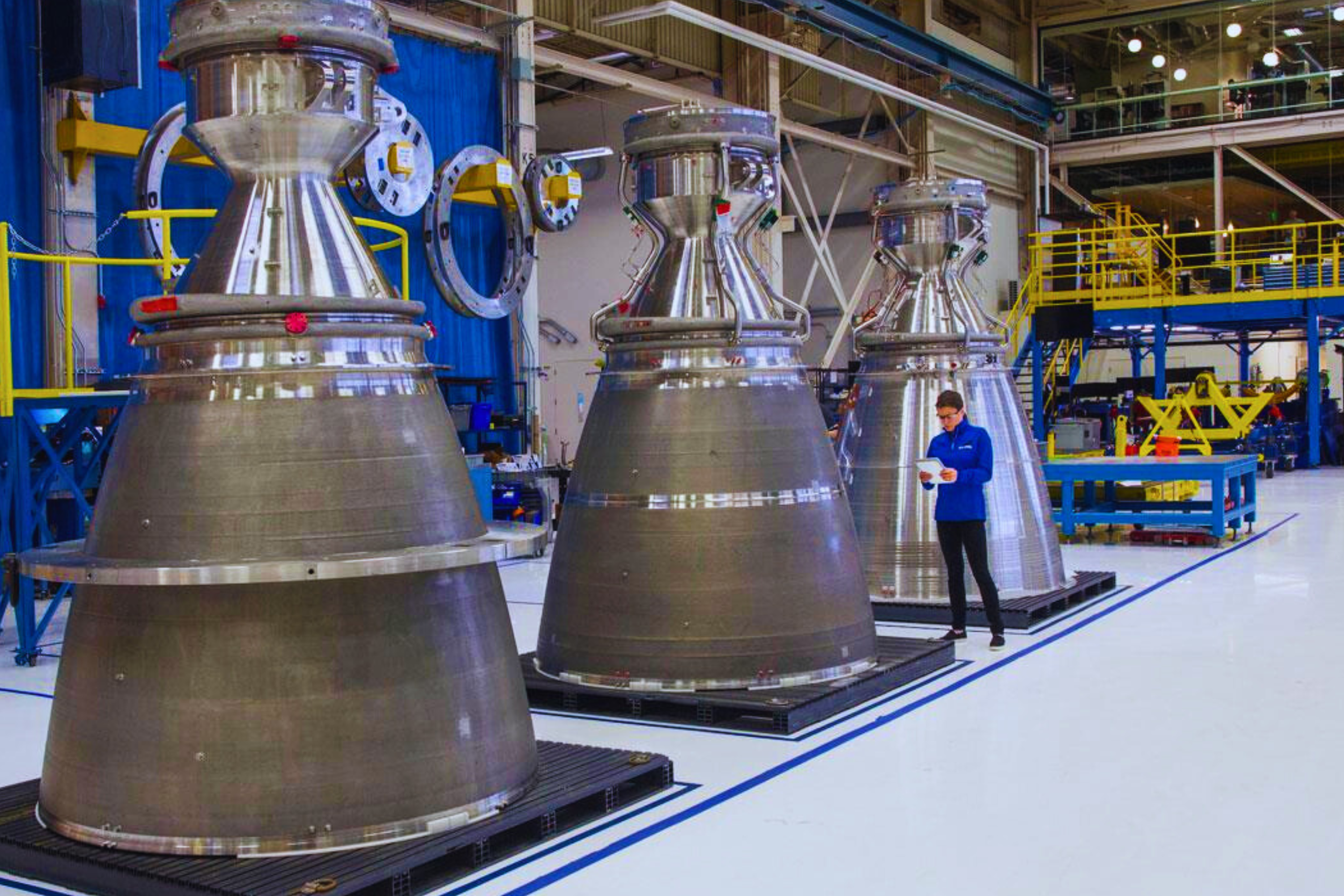 Three large rocket engine components in a manufacturing facility, with a person inspecting one of them.