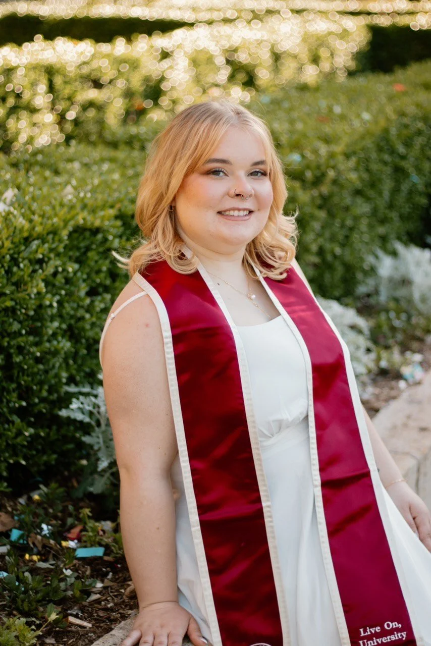 Young woman with blonde hair wearing a white dress and maroon graduation stole outdoors, smiling at the camera.