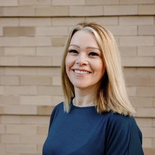 A smiling woman with blonde hair, wearing a navy blue top, standing in front of a beige brick wall.