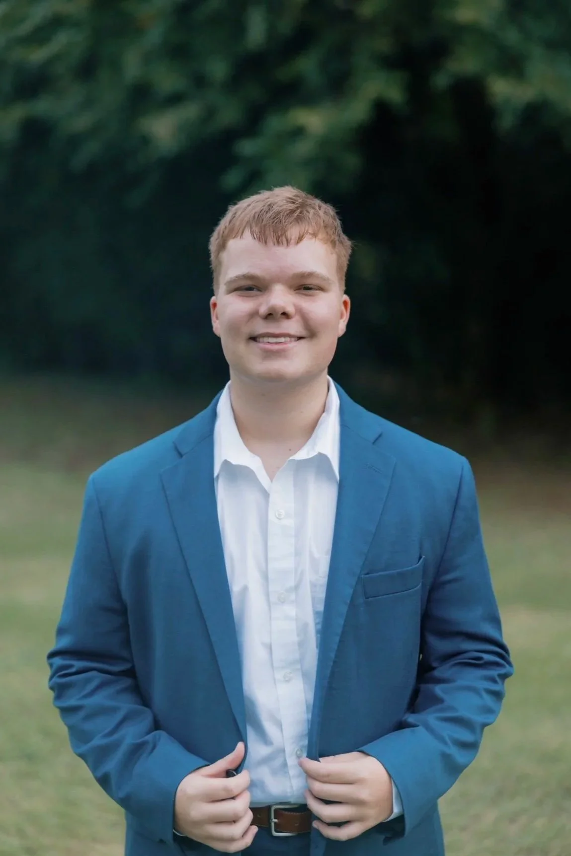 A young man smiling outdoors, wearing a blue blazer and white shirt, standing on a grassy area with trees in the background.