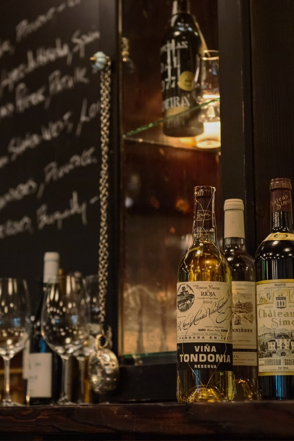 Wine bottles and glasses on a bar with a chalkboard in the background.