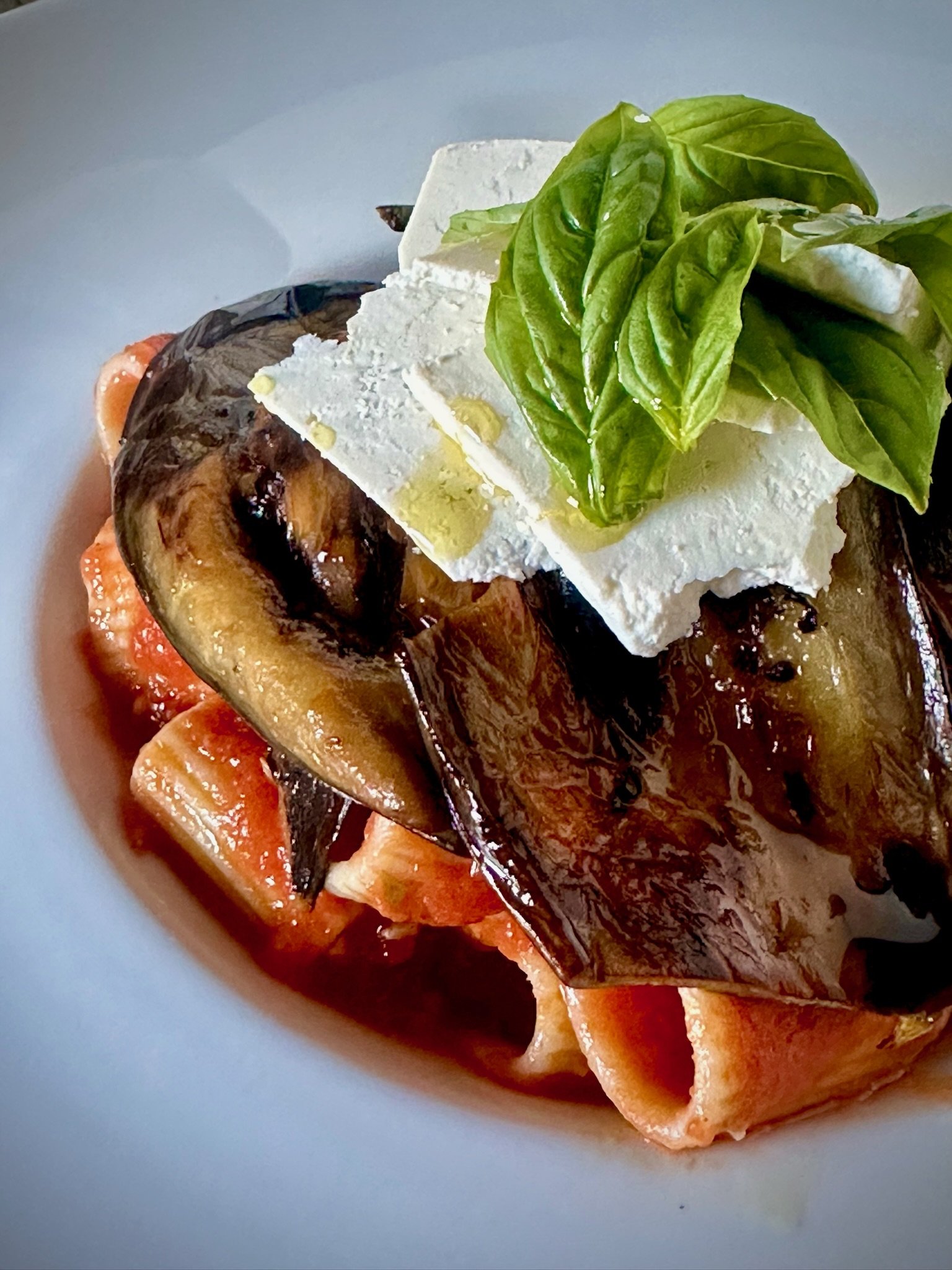 Pasta dish with rigatoni topped with grilled eggplant slices, ricotta cheese, fresh basil leaves, and a drizzle of olive oil on a white plate.
