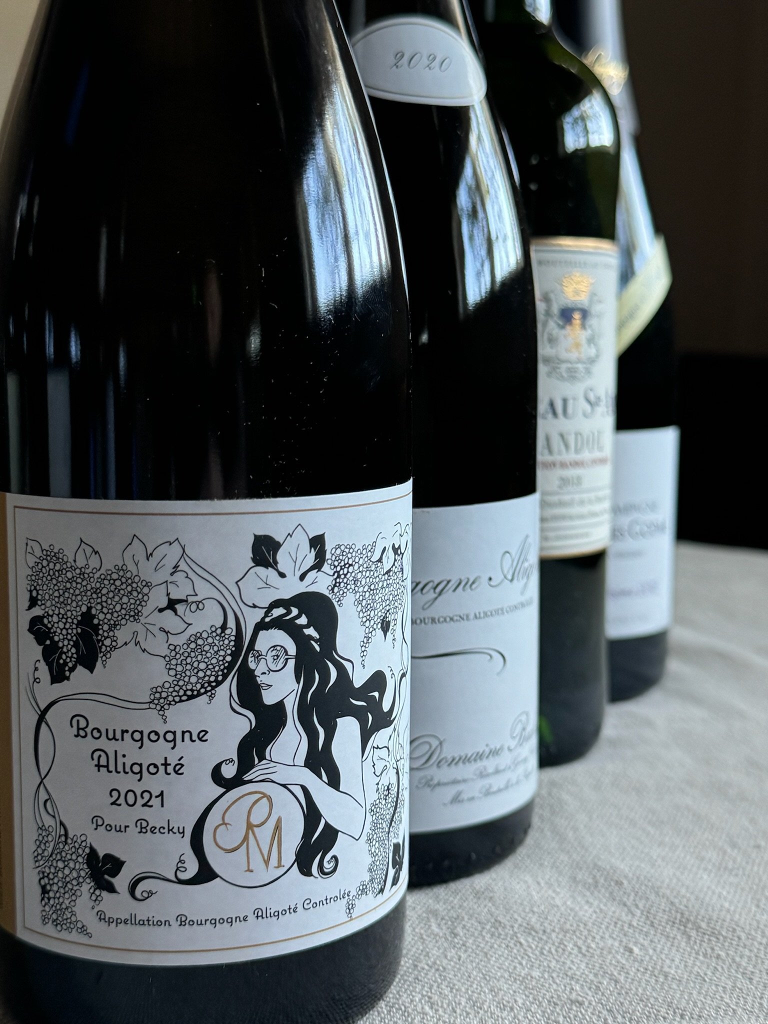 Four wine bottles on a table, including Bourgogne Aligoté 2021 with an artistic black and white label featuring a woman and grape vines.