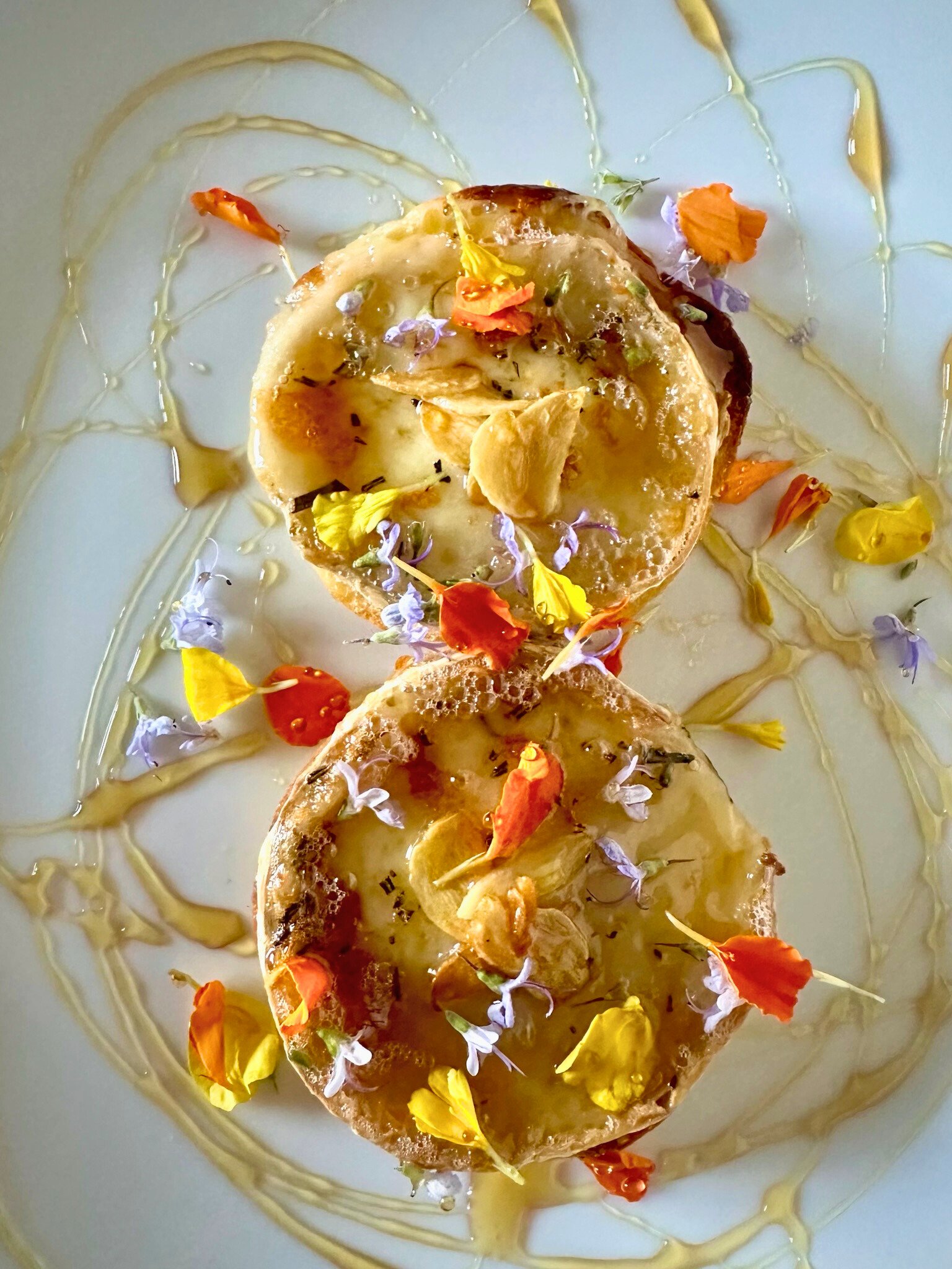 Two gourmet pancakes garnished with honey drizzle and colorful edible flowers on a white plate.