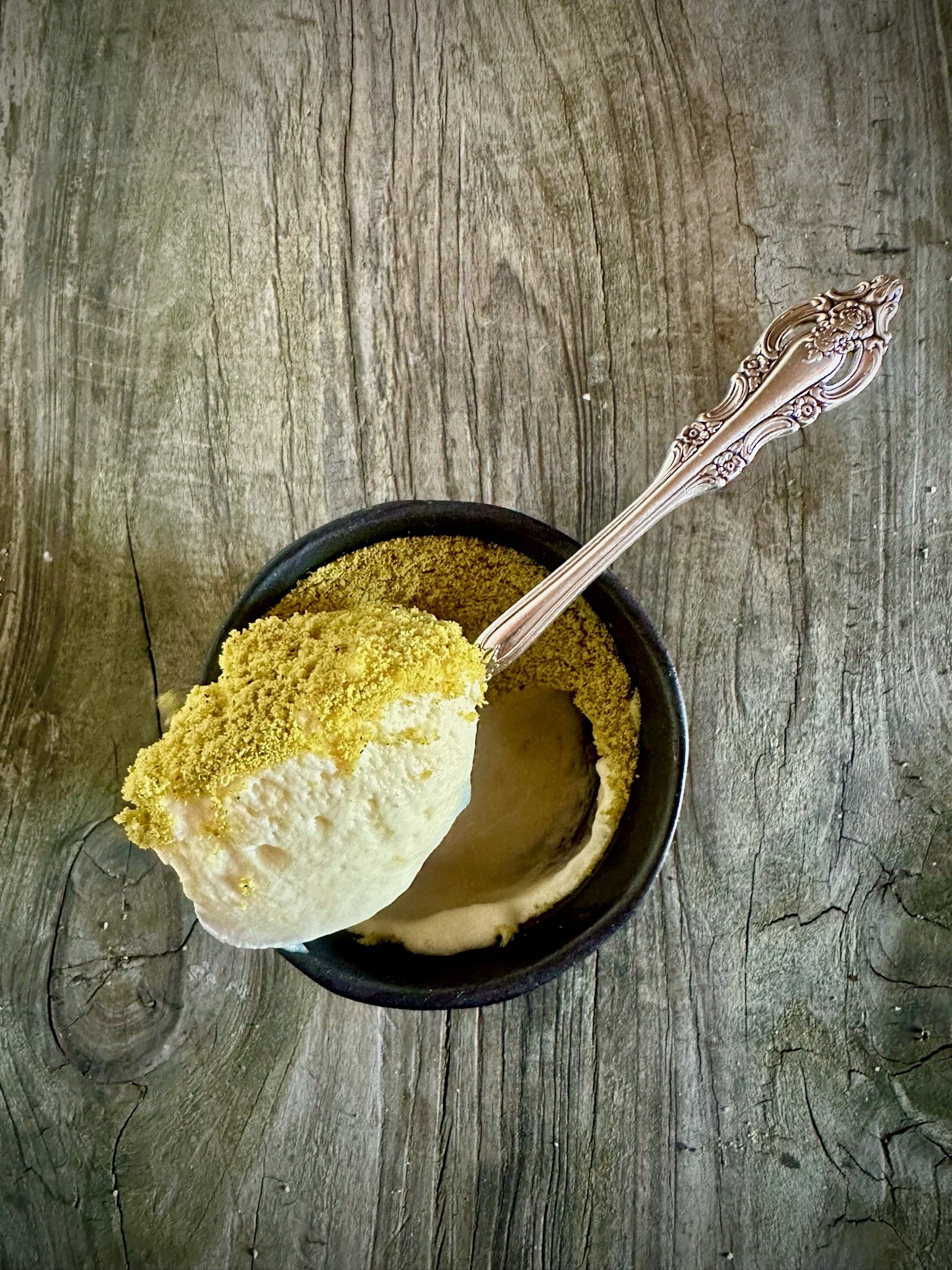 A bowl of pistachio ice cream on a wooden surface, with a scoop on an ornate spoon.