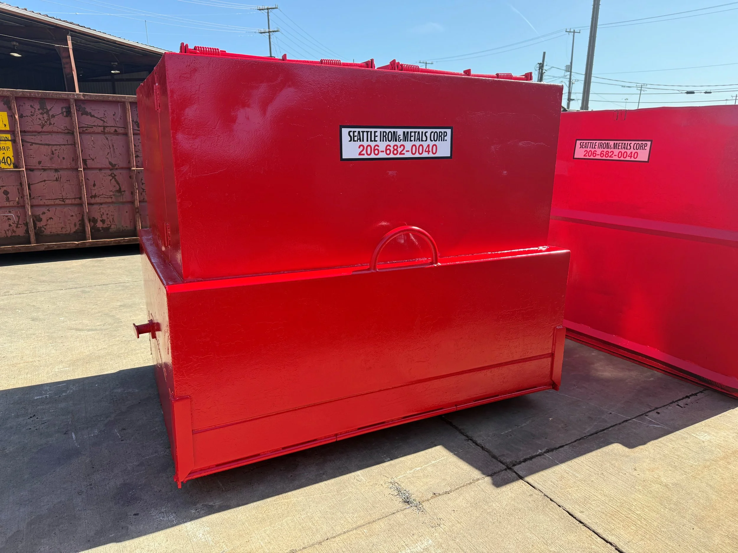 Red metal container with a label from Seattle Iron & Metals Corp, positioned outdoors on a concrete surface, with other containers in the background and power lines visible in the sky.