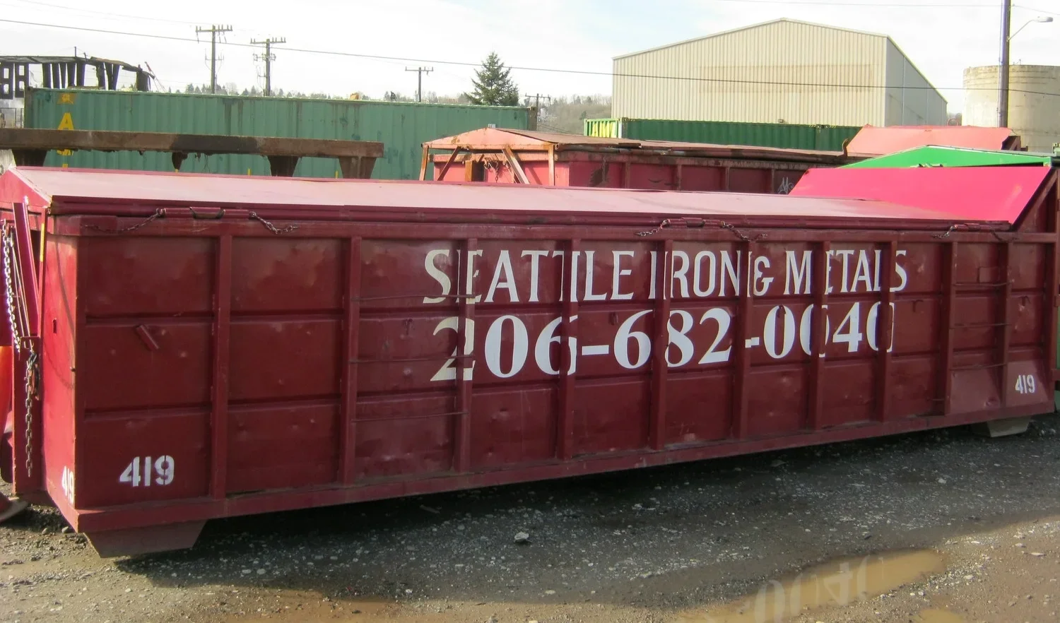 Red metal dumpster with white stenciled lettering reading 'SEATTLE IRON & METALS 206-682-0640' and the number 419 in the lower left corner, located outdoors on a gravel ground with industrial buildings and containers in the background.