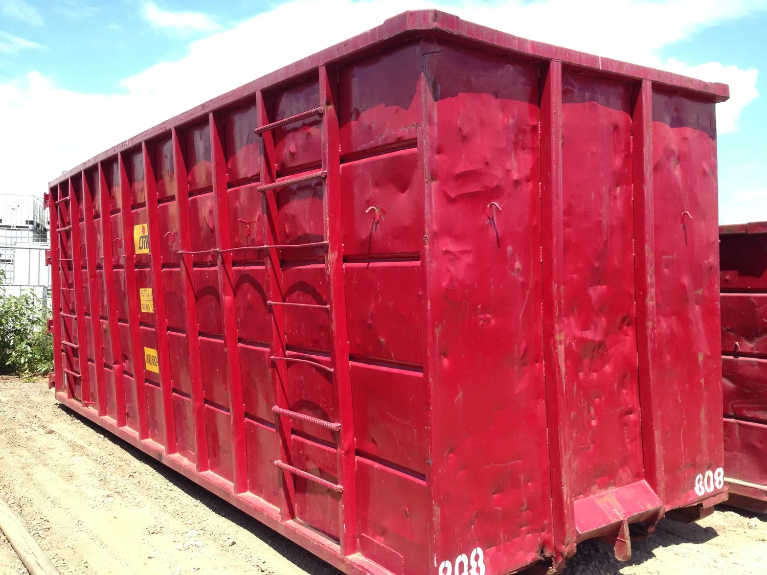Red industrial dumpster with yellow labels on a gravel surface under a partly cloudy sky.