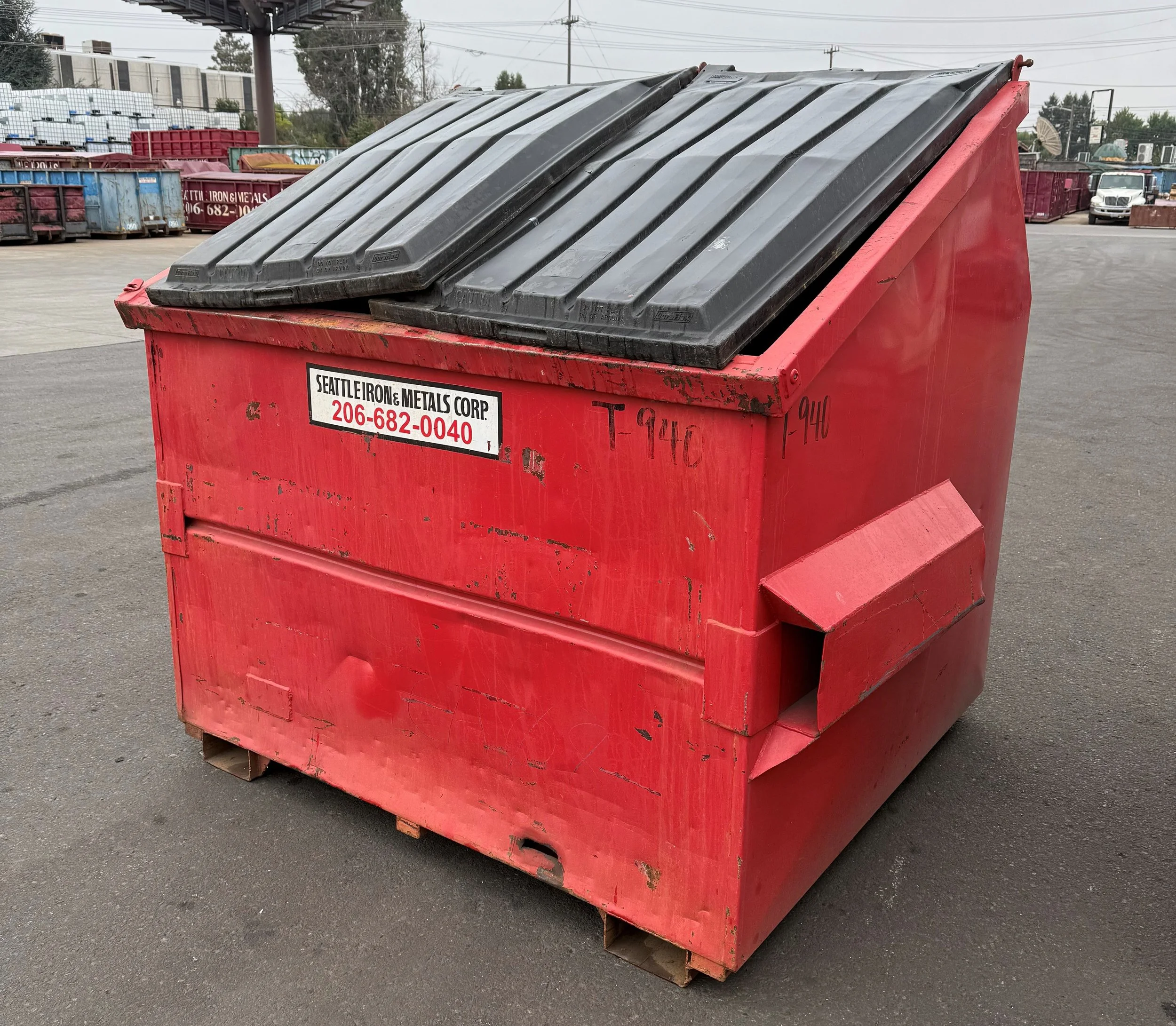 A red industrial dumpster with black lids and a sign for Seattle Iron & Metals Corp in an outdoor lot with other dumpsters and trucks in the background.