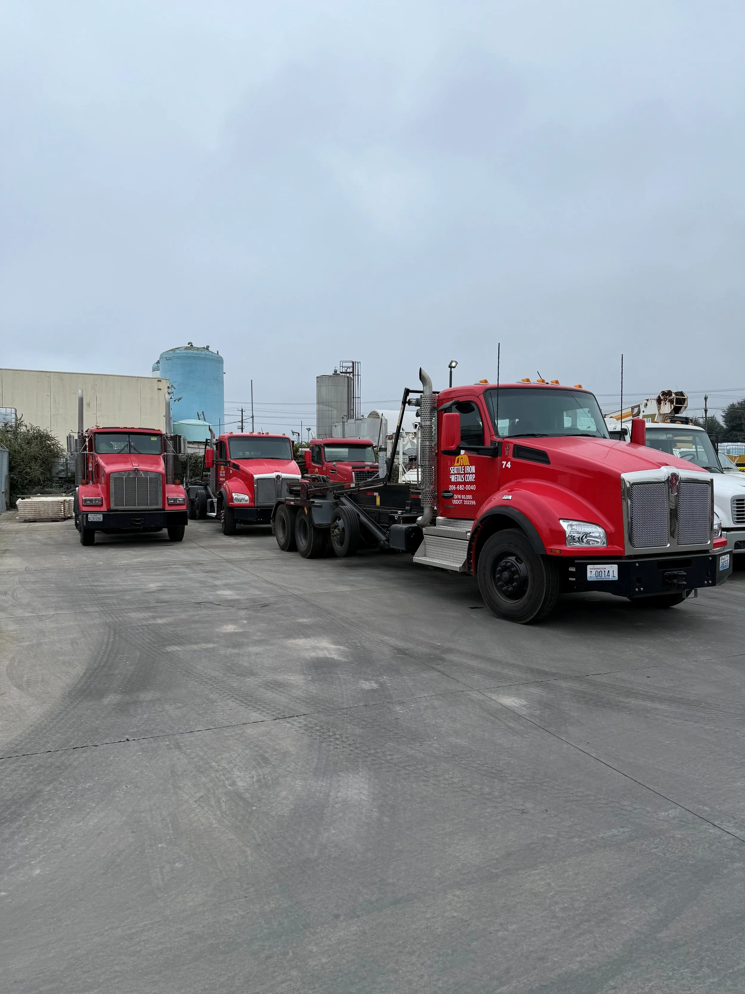 A row of red semi-trucks parked on a concrete lot with industrial buildings and tanks in the background.