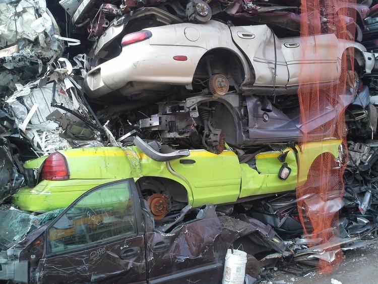 Pile of crushed and mangled cars stacked on top of each other, appearing to be at a junkyard or scrap yard.