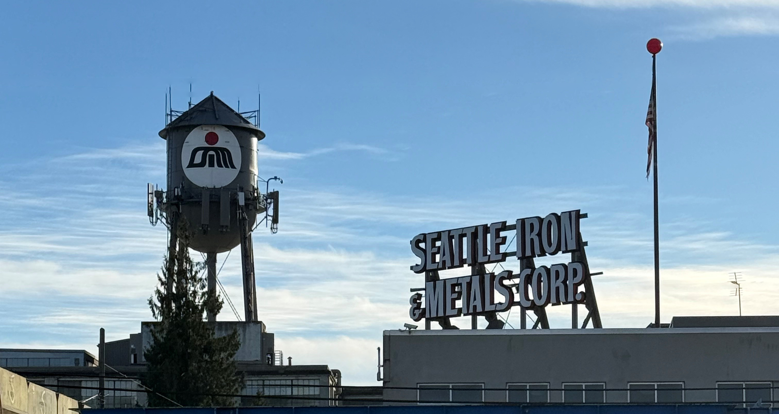 Sign reading 'Seattle Iron & Metals Corp' next to a flagpole with an American flag and a red ball on top, with a historic water tower bearing a logo against a blue sky.