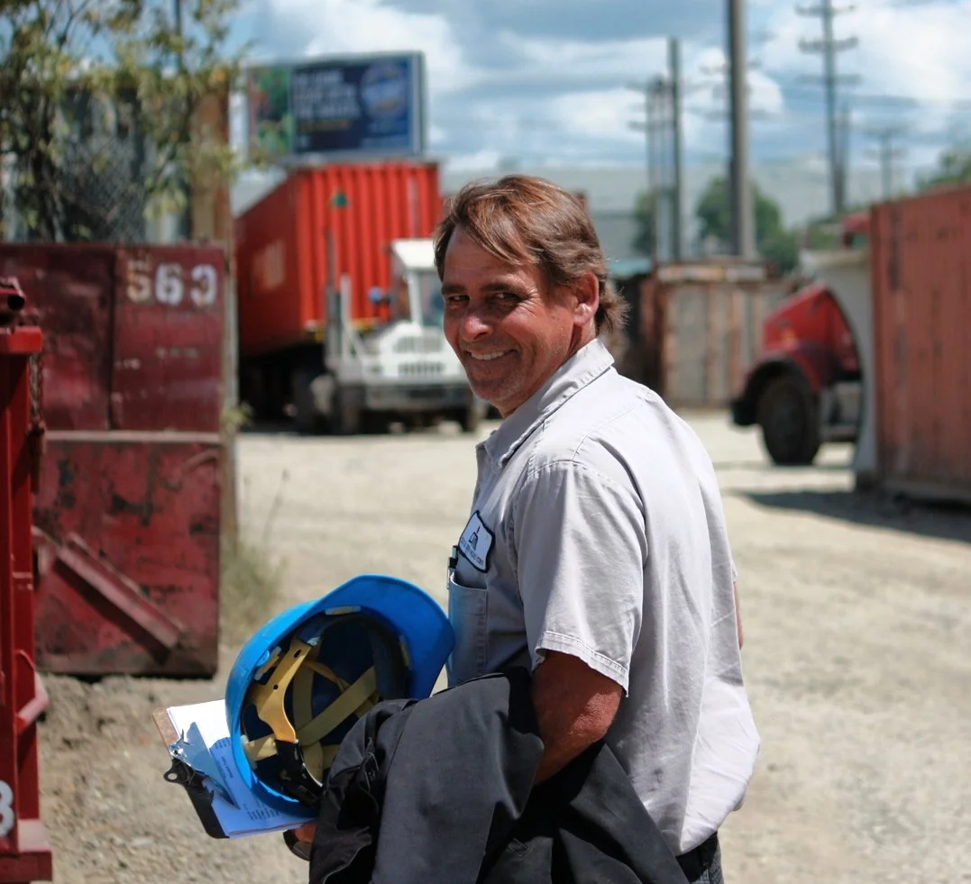 A man in a white shirt holding a blue helmet and black jacket, smiling outdoors near red and orange trucks with a blurry background of a parking lot and power lines.