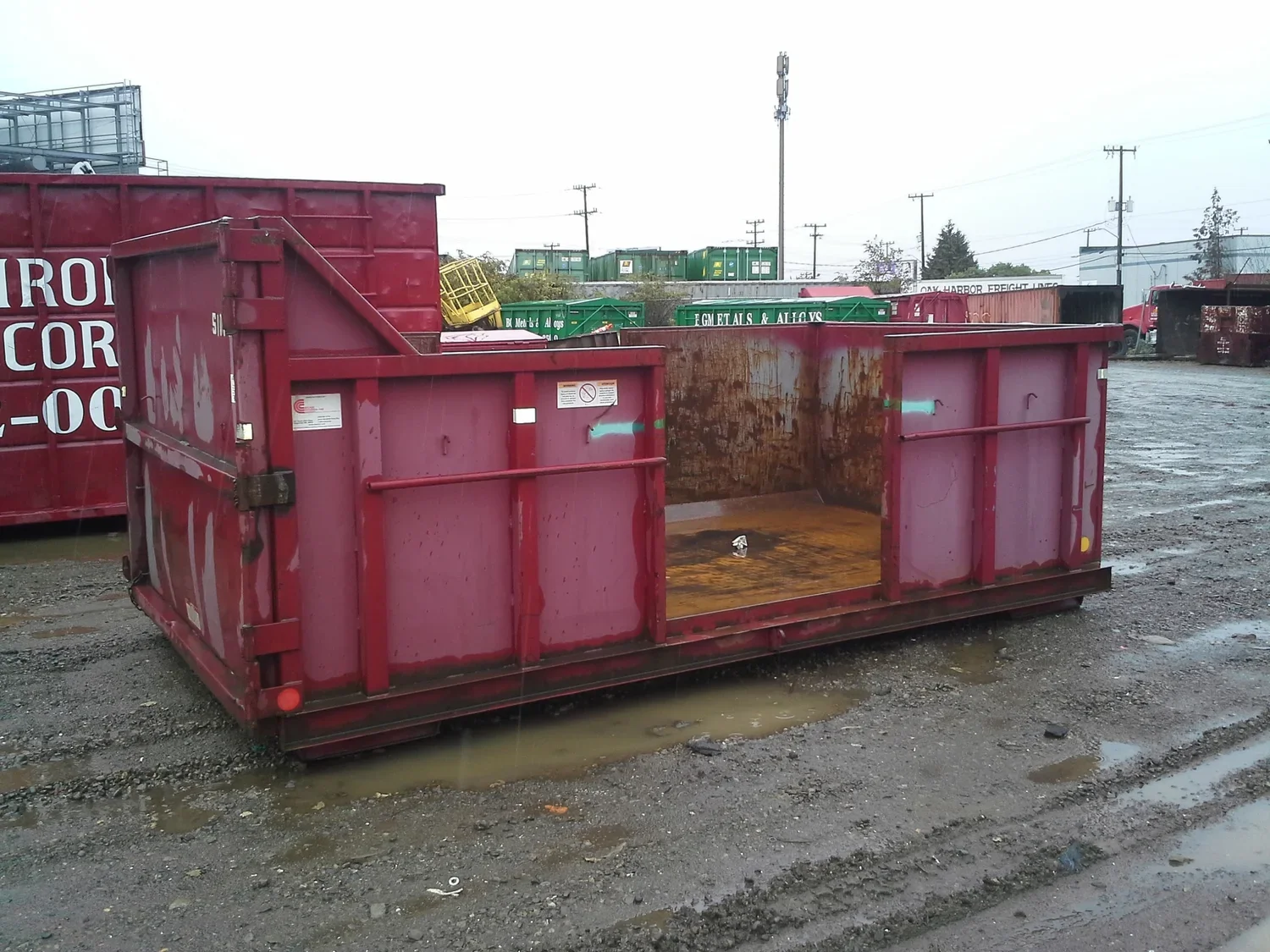An empty red industrial dumpster on a muddy, wet ground in a lot with other dumpsters and train cars in the background.