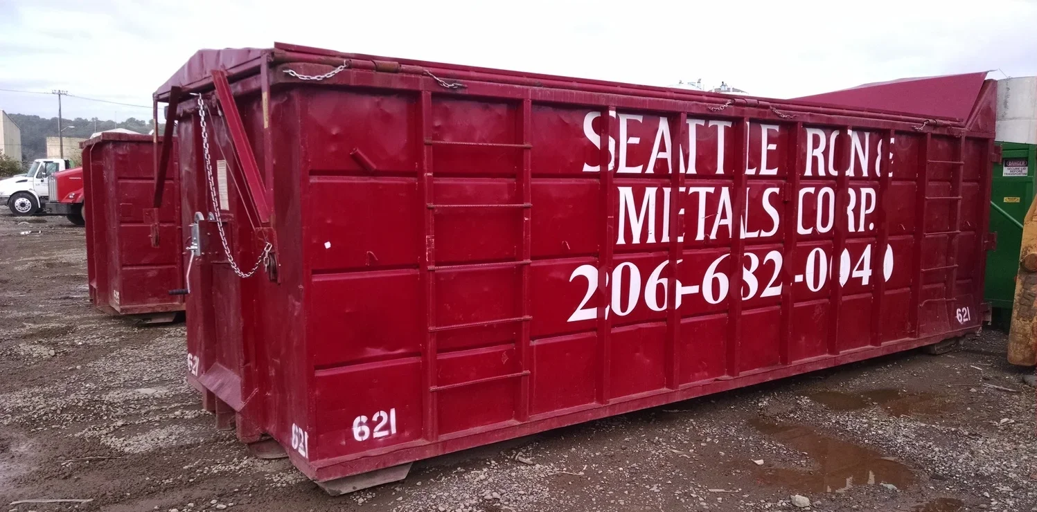 Large red dumpster with white lettering that reads 'Seattle Rone Metals Corp.' along with a phone number, placed on a gravel lot with cars and other equipment in the background.