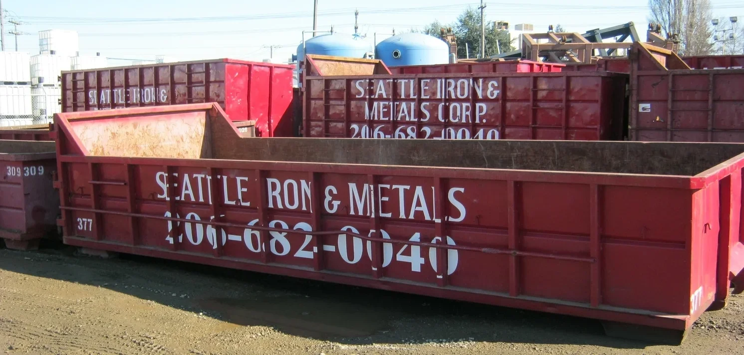 Red open-top metal shipping container with white painted text that reads 'SEATTLE IRON & METALS 206-682-0046'.