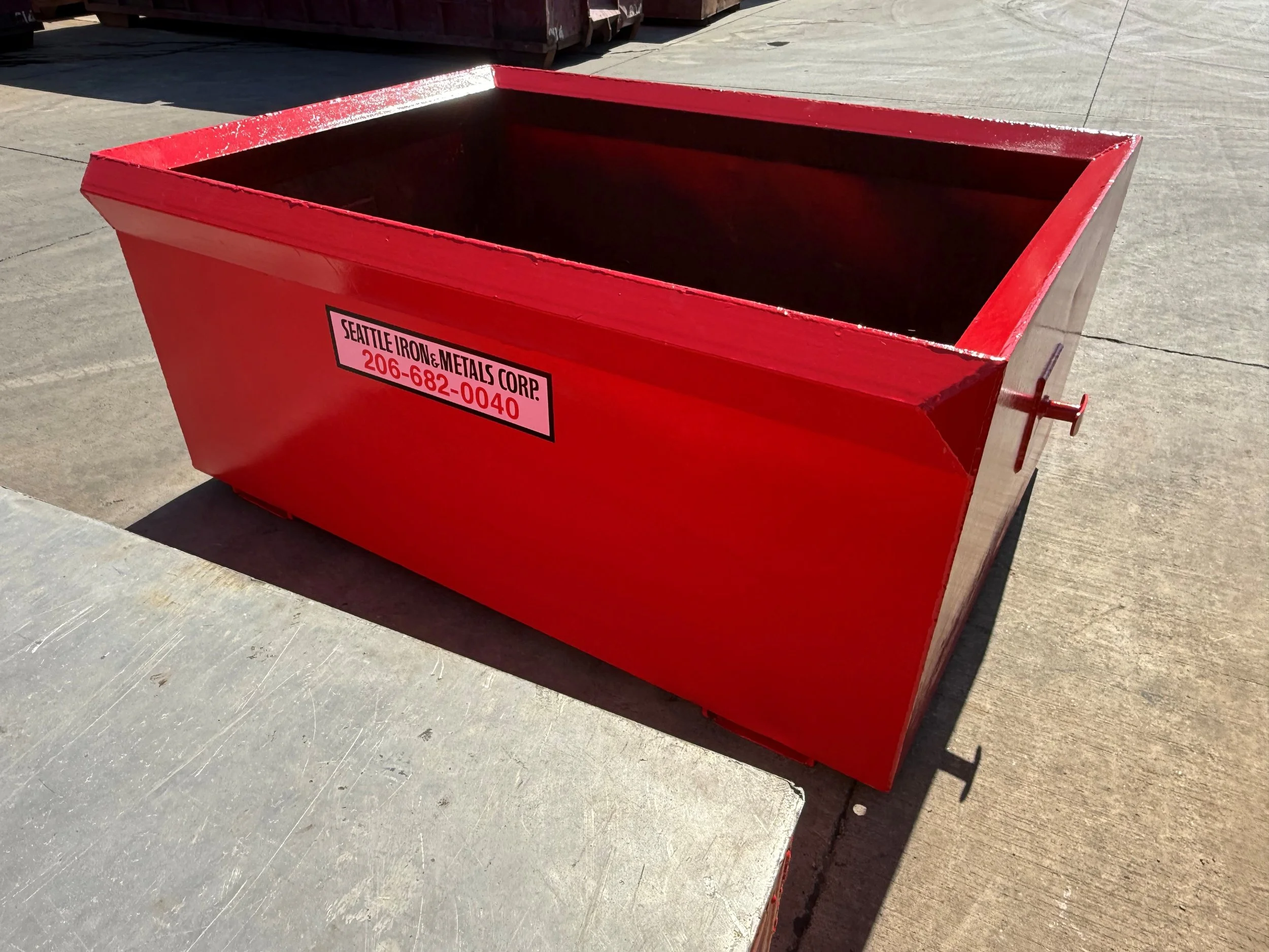 Large red metal delivery tub with a label for Seattle Iron & Metals Corp, placed on concrete pavement.