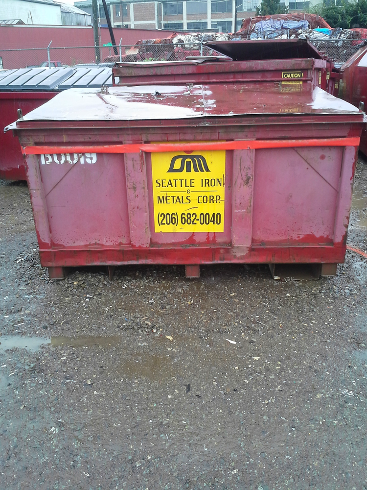 Red dumpster with yellow sign reading 'Seattle Iron & Metals Corp' and phone number, situated on a gravel lot with other containers and industrial background.
