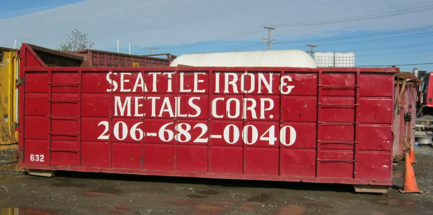 Red construction dumpster with white text that reads "Seattle Iron & Metals Corp." and a phone number, parked outdoors with an orange traffic cone nearby.