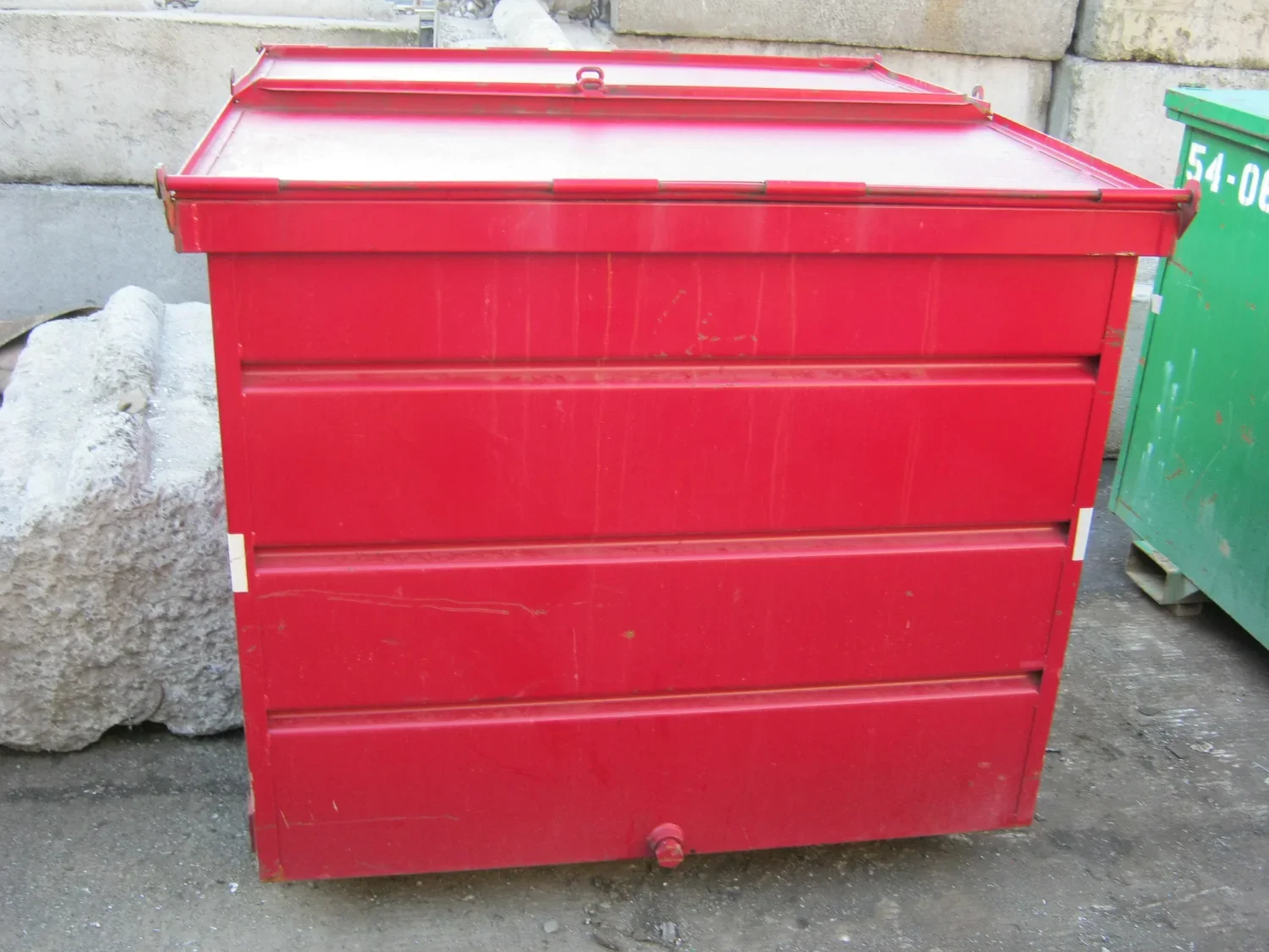 A red metal collection bin outdoors on a concrete surface, with a green container partially visible to the right and a large rock to the left.