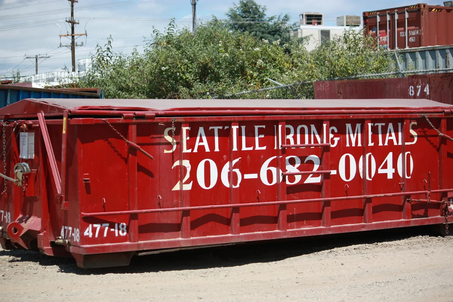 Red steel container with white lettering that says 'Seattle Iron & Metals' along with a phone number, placed on a dirt ground outdoors with shipping containers and electric poles in the background.