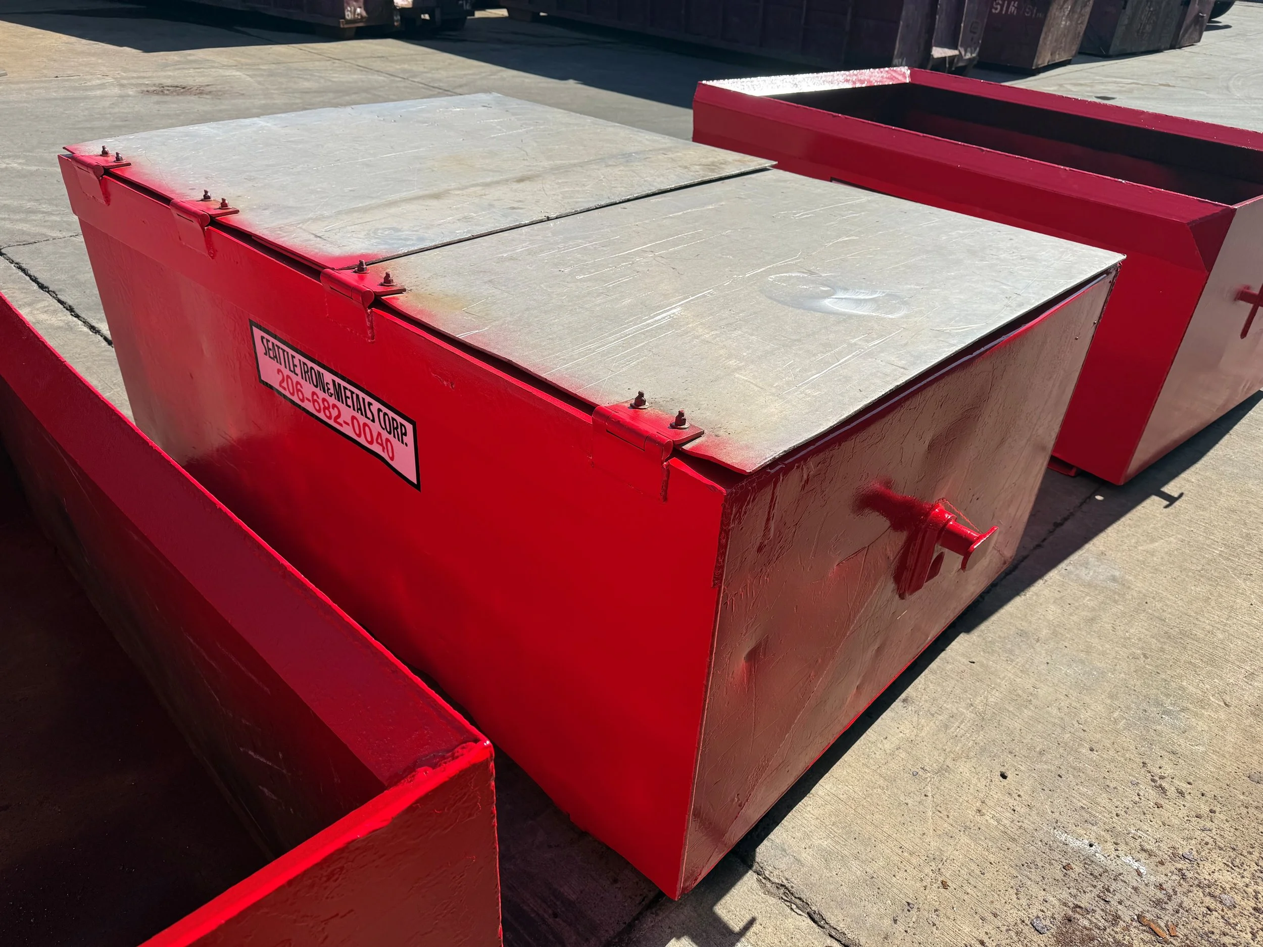 Large red metal container with a silver cover secured with bolts, labeled 'Seattle Iron & Metals Corp' with a phone number.