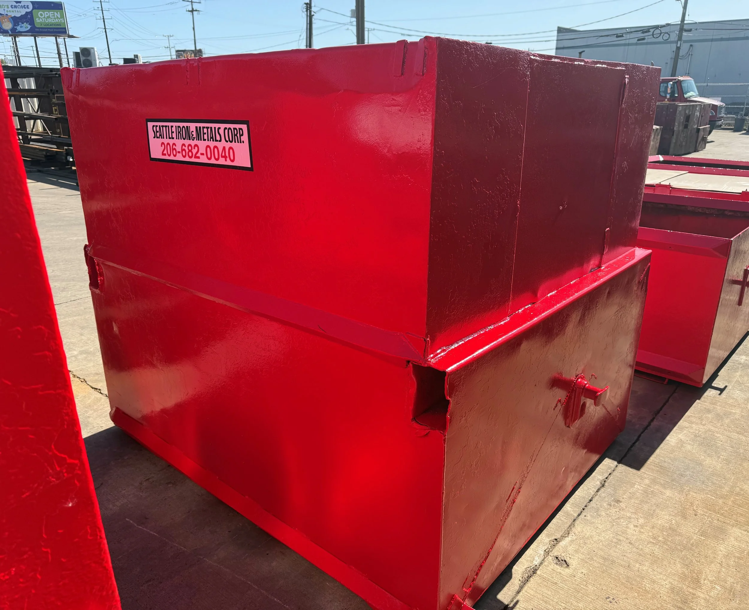 Large red metal container with a black and pink label that reads 'Seattle Iron & Metals Corp. 206-682-0040' placed outdoors on a concrete surface, with other metal containers and trucks visible in the background.