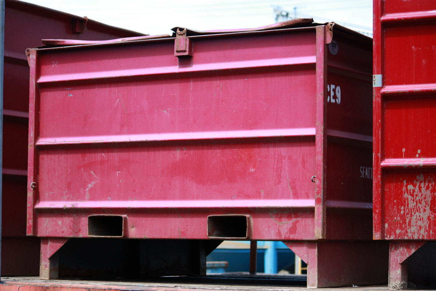 Close-up of a red shipping container with visible wear and scratches, partially stacked with other containers in the background.
