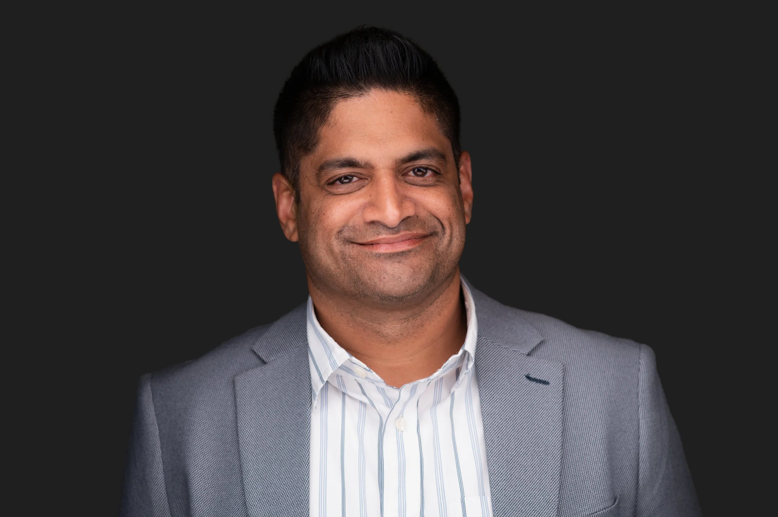 A professional portrait of a man with short dark hair, smiling, wearing a light gray suit jacket and a white striped shirt, against a dark background.