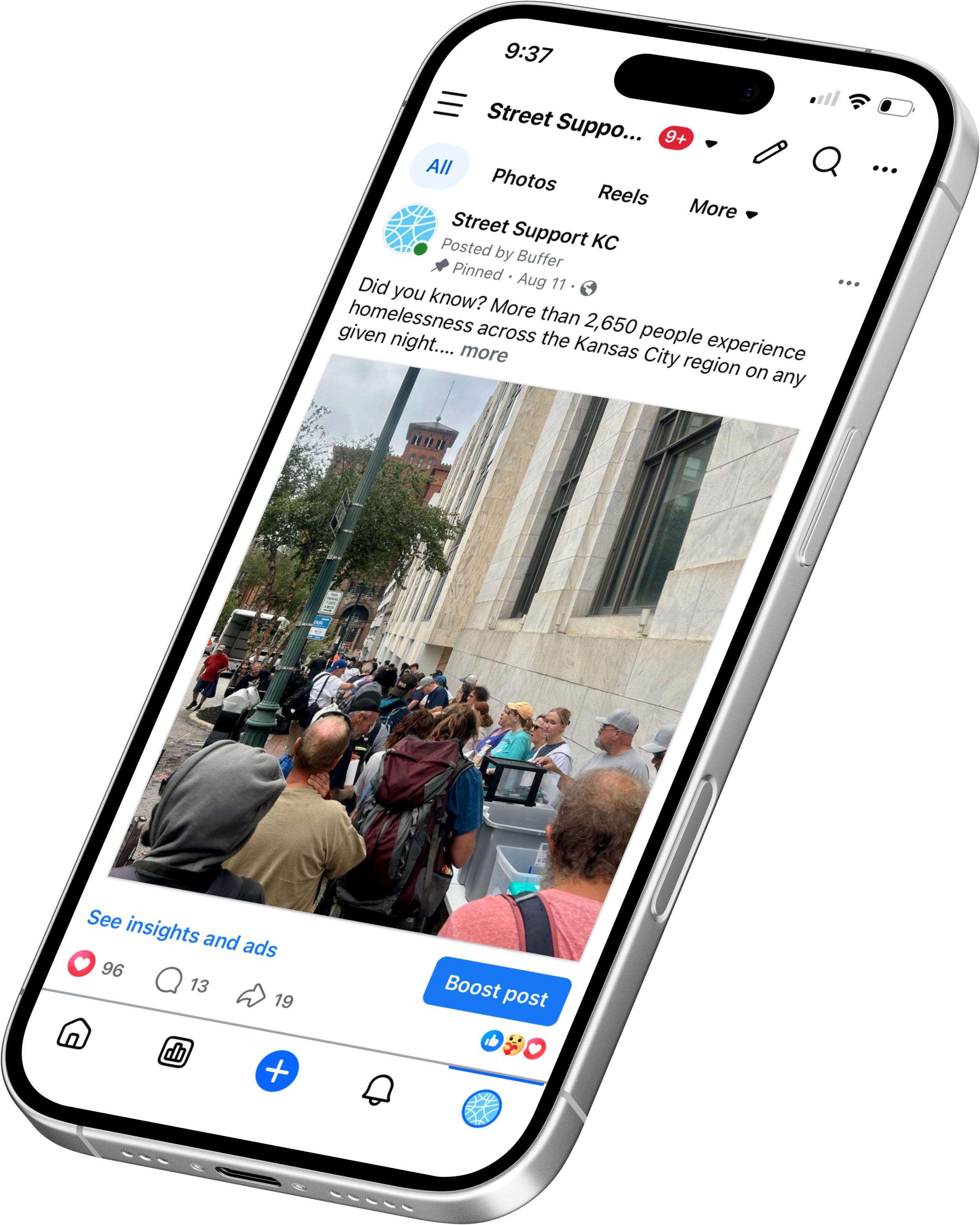 A smartphone screen displaying a Facebook post from Street Support KC. The post shares a fact about homelessness in Kansas City, with a partially visible photo showing a line of people waiting outside a building on a city street.