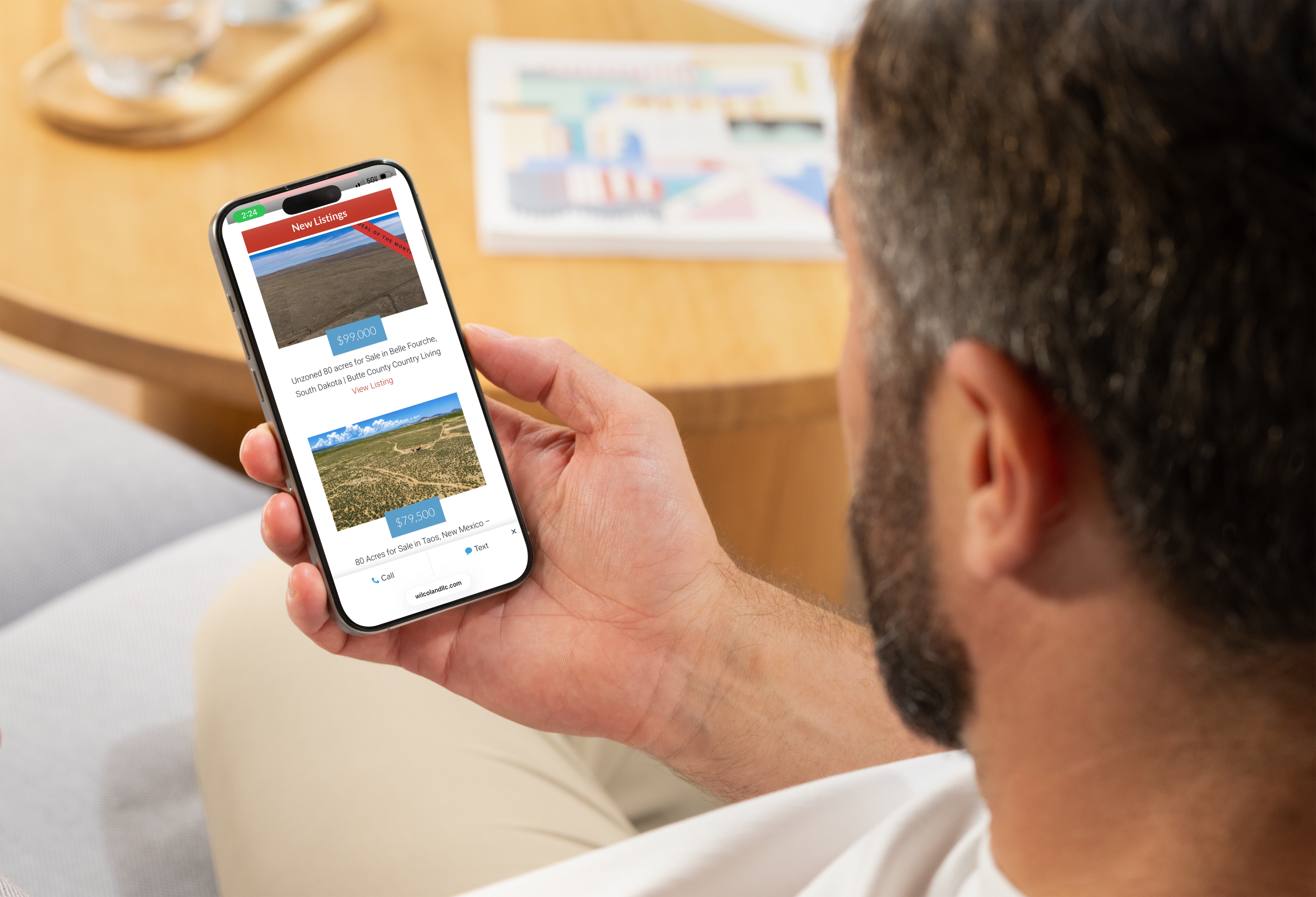 A person holding a smartphone displaying a real estate listing app, with land for sale in South Dakota and New Mexico. The person has short curly hair and is wearing a white shirt, seated at a wooden table with brochures in the background.