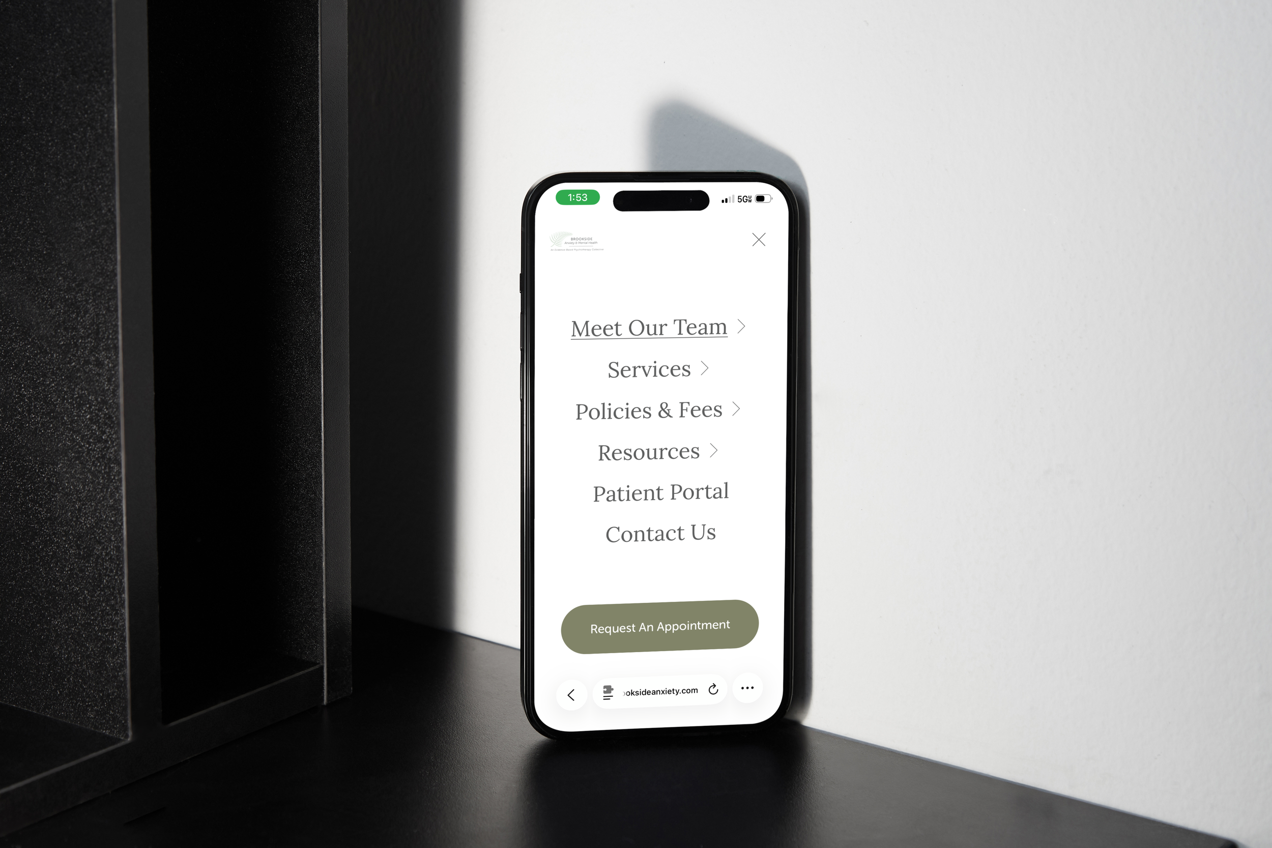 Smartphone on a black surface displaying a website with sections titled 'Meet Our Team,' 'Services,' 'Policies & Fees,' 'Resources,' 'Patient Portal,' 'Contact Us,' and a green button labeled 'Request An Appointment.'