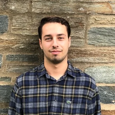 Emmy Award winning producer Jesse Seidman stands in front of an aged stone building in a flannel shirt with a slight smile to smirk on his face