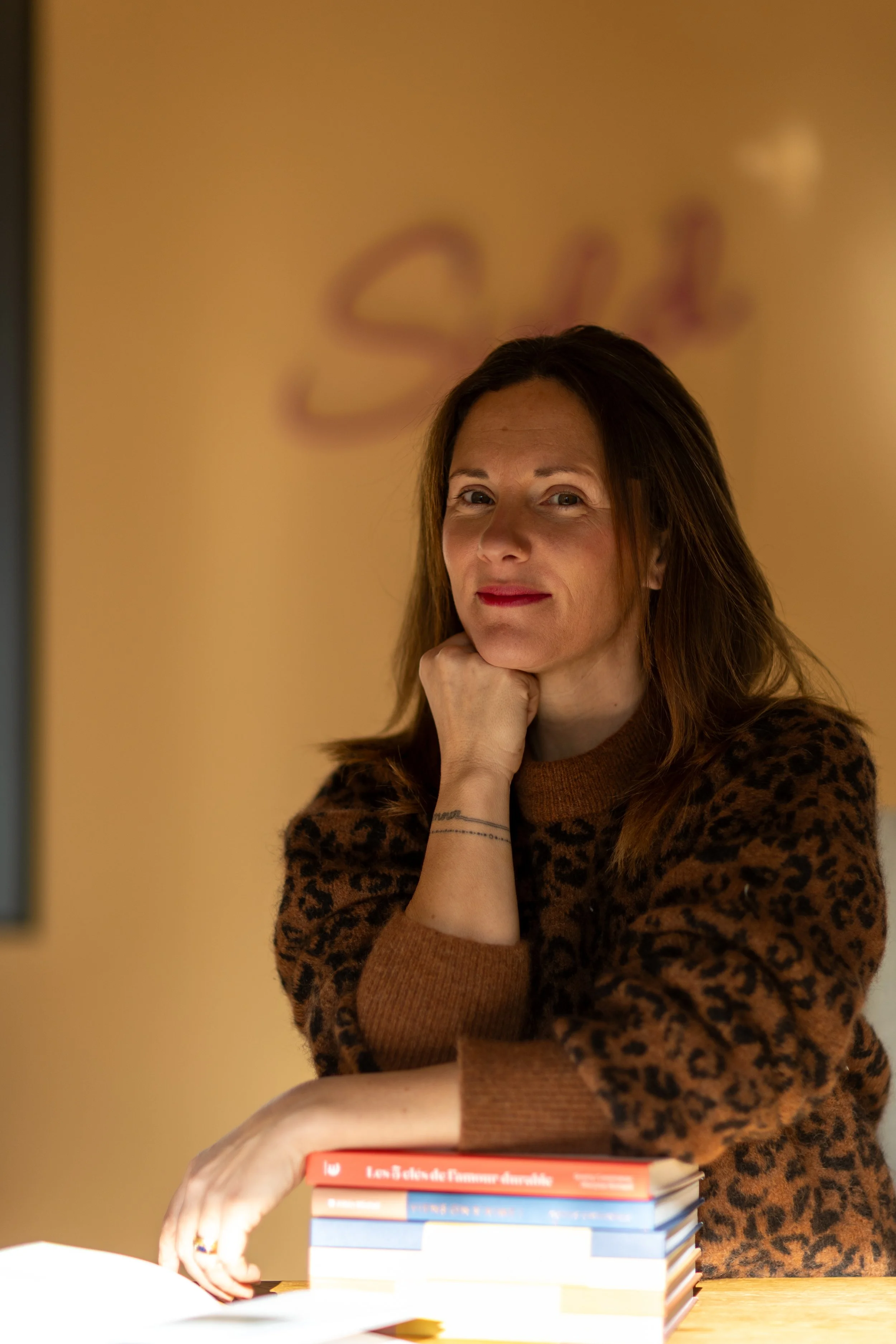 Une femme avec des cheveux bruns, portant un pull à motif léopard marron, posant avec ses bras reposant sur une pile de livres, dans un environnement intérieur chaleureux.