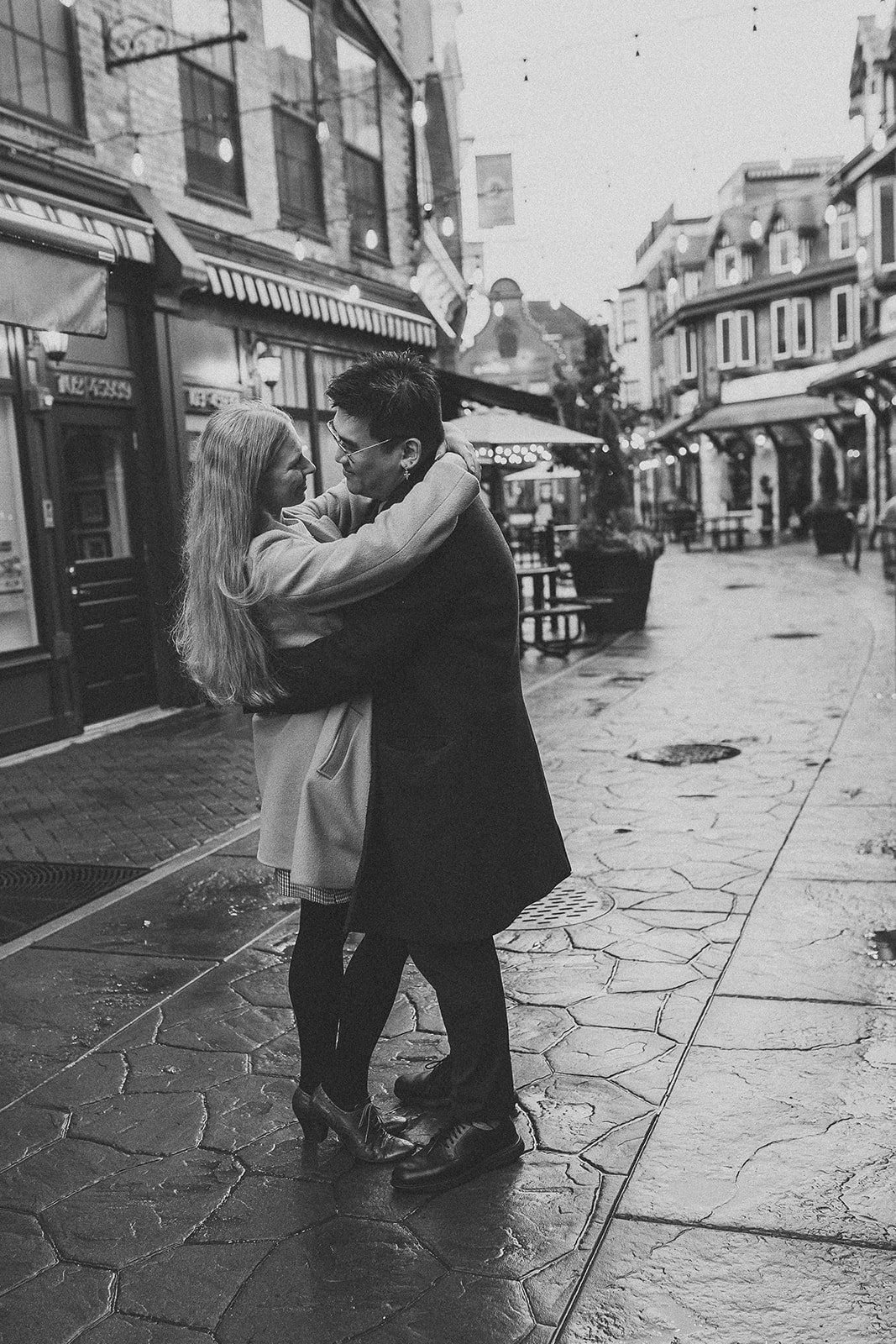 A black and white photo of a couple embracing on a cobblestone street in a downtown area with shops and outdoor seating.