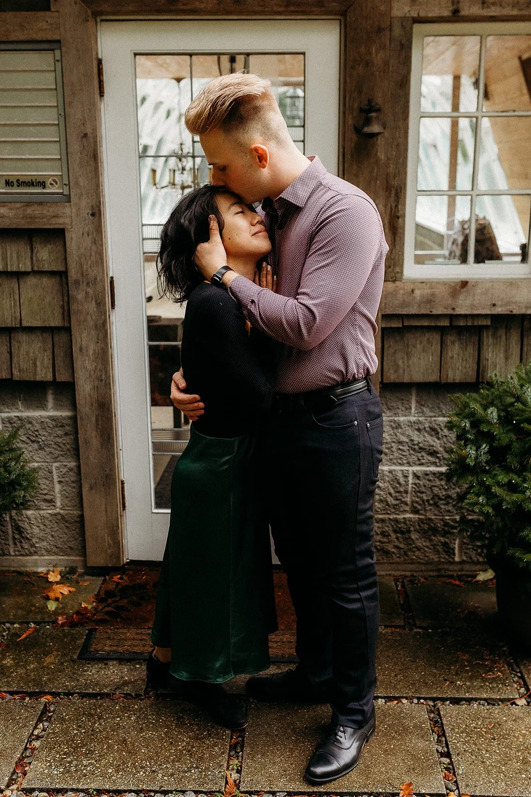 A couple hugging outside a rustic building with wooden siding. The man is kissing the woman's forehead while holding her face, and the woman is smiling with her eyes closed. There are small trees in pots on either side of the couple, and fallen leave