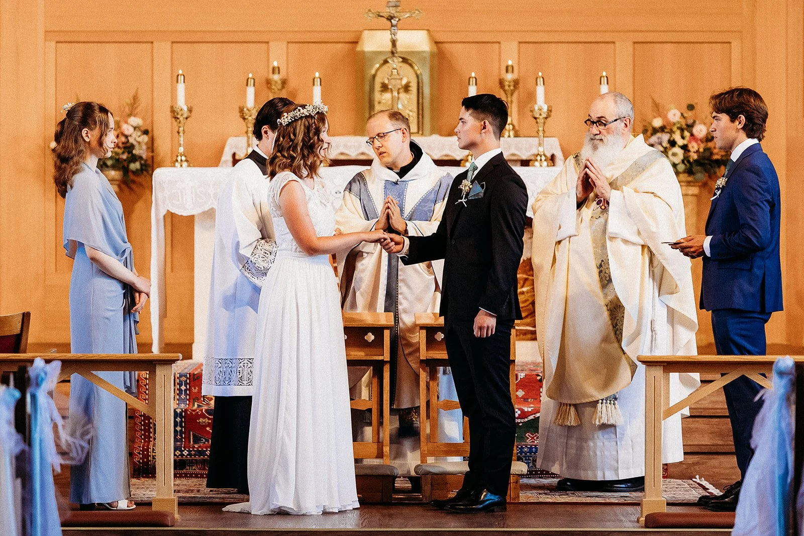 A wedding ceremony inside a church, with the bride and groom holding hands and facing each other, surrounded by the officiant, bridesmaid, and groomsman, with an altar in the background.
