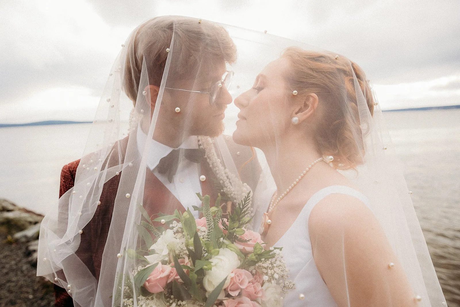 A bride and groom standing close together under a wedding veil, near a body of water on a cloudy day. The bride holds a bouquet of pink and white flowers, and both are wearing pearl jewelry.