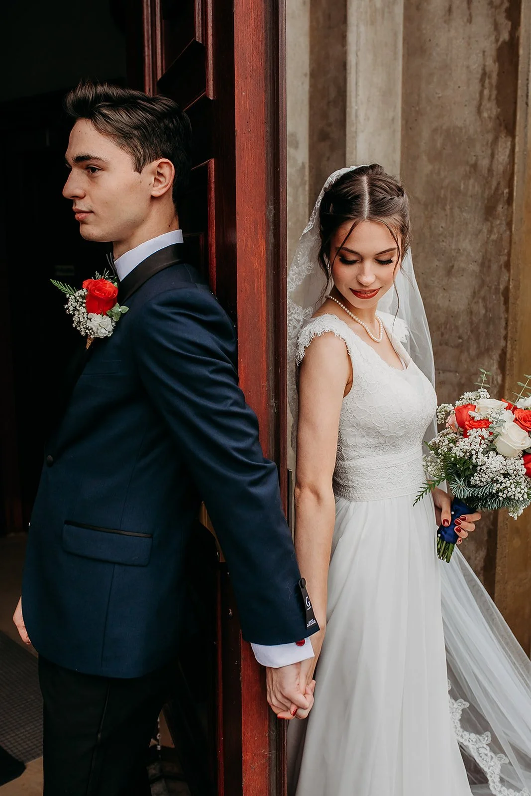 A bride and groom standing back to back separated by a doorframe, holding hands. The groom is in a dark suit with a boutonniere, and the bride is in a white lace wedding dress, holding a bouquet of red and white flowers.