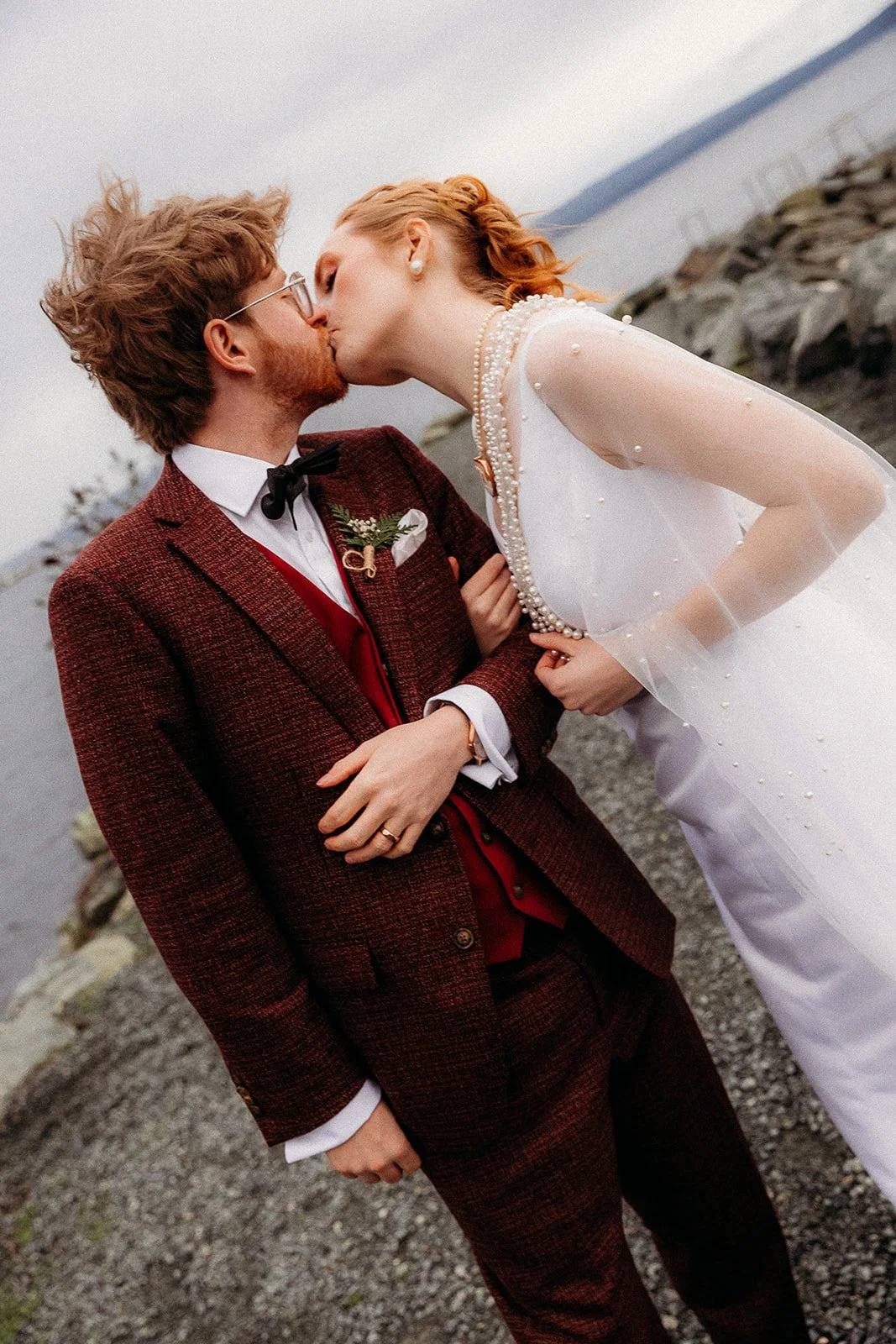 A couple dressed in wedding attire sharing a kiss outdoors near a body of water and rocks.