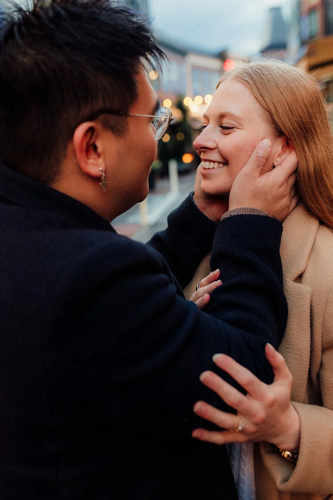 A couple sharing a tender moment on a city street at dusk, with the woman smiling as the man gently holds her face.