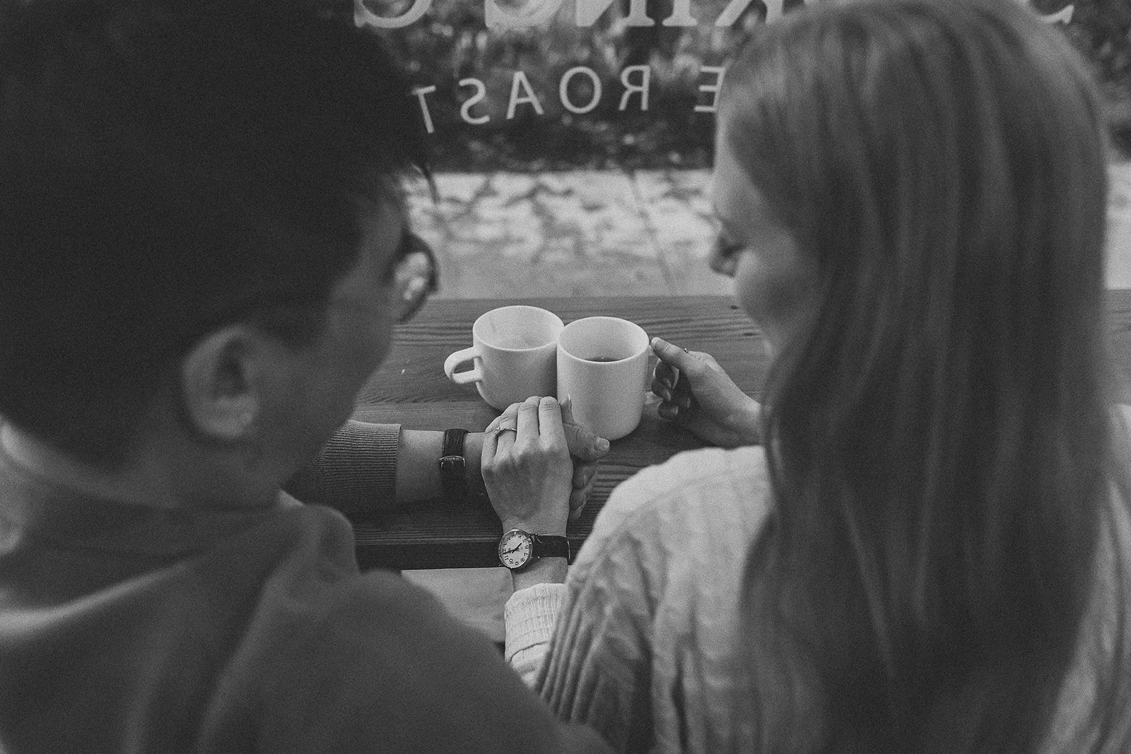 A black and white photo of a man and woman sitting at a wooden table, holding hands, with coffee mugs in front of them, in a cozy cafe setting.